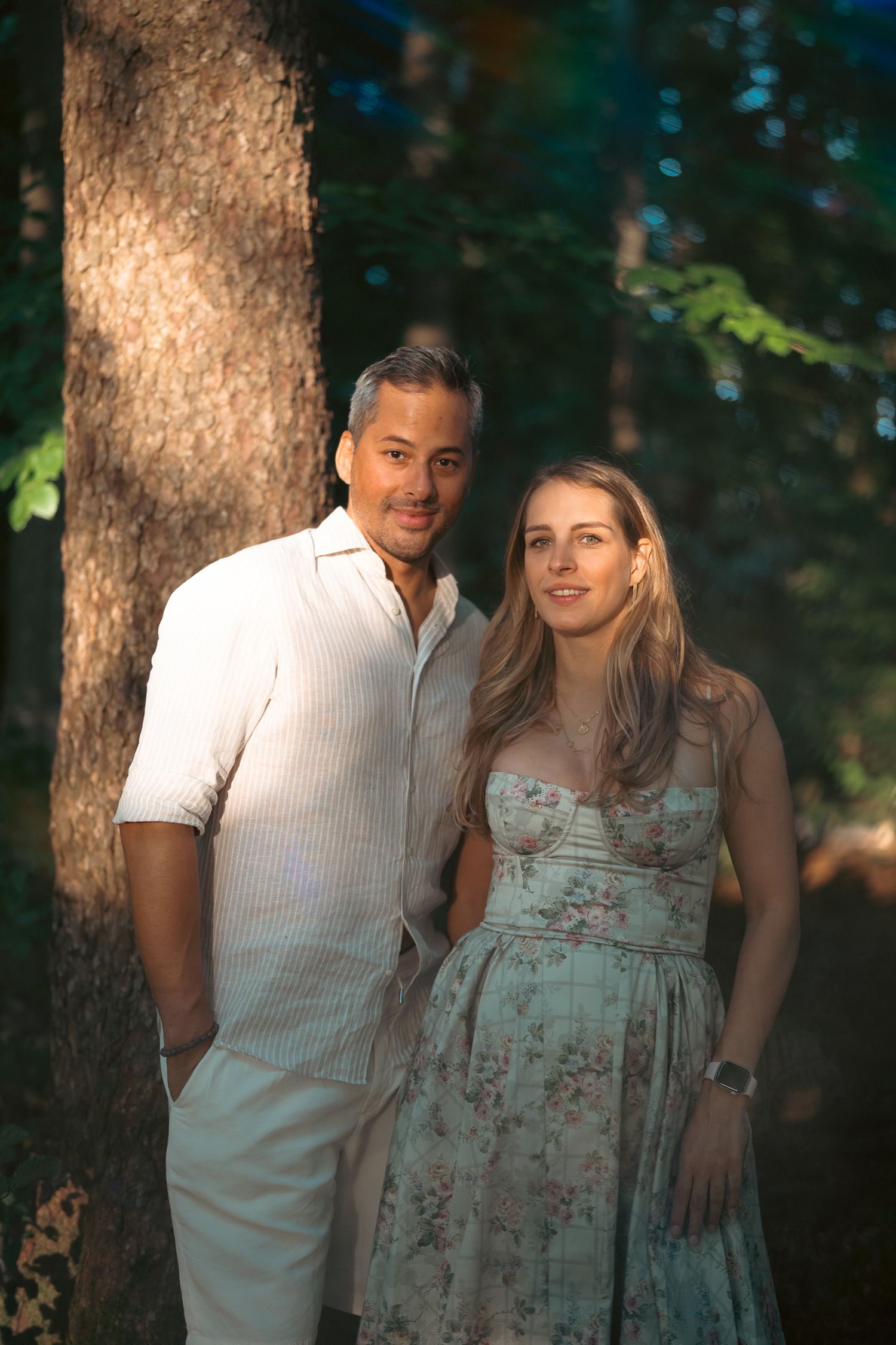 Couple posing together in a sunlit forest, man in white shirt and woman in floral dress smiling.