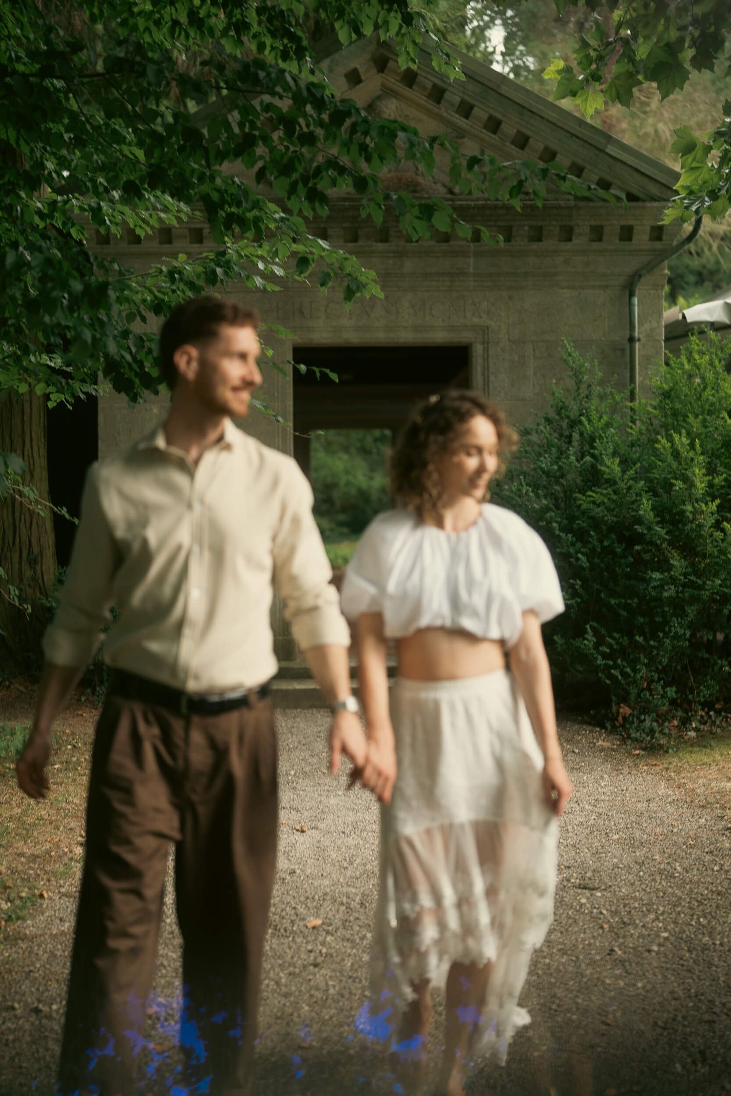 A couple holds hands while walking outdoors near a rustic building surrounded by lush green trees. Villa Boveri Wedding, Baden