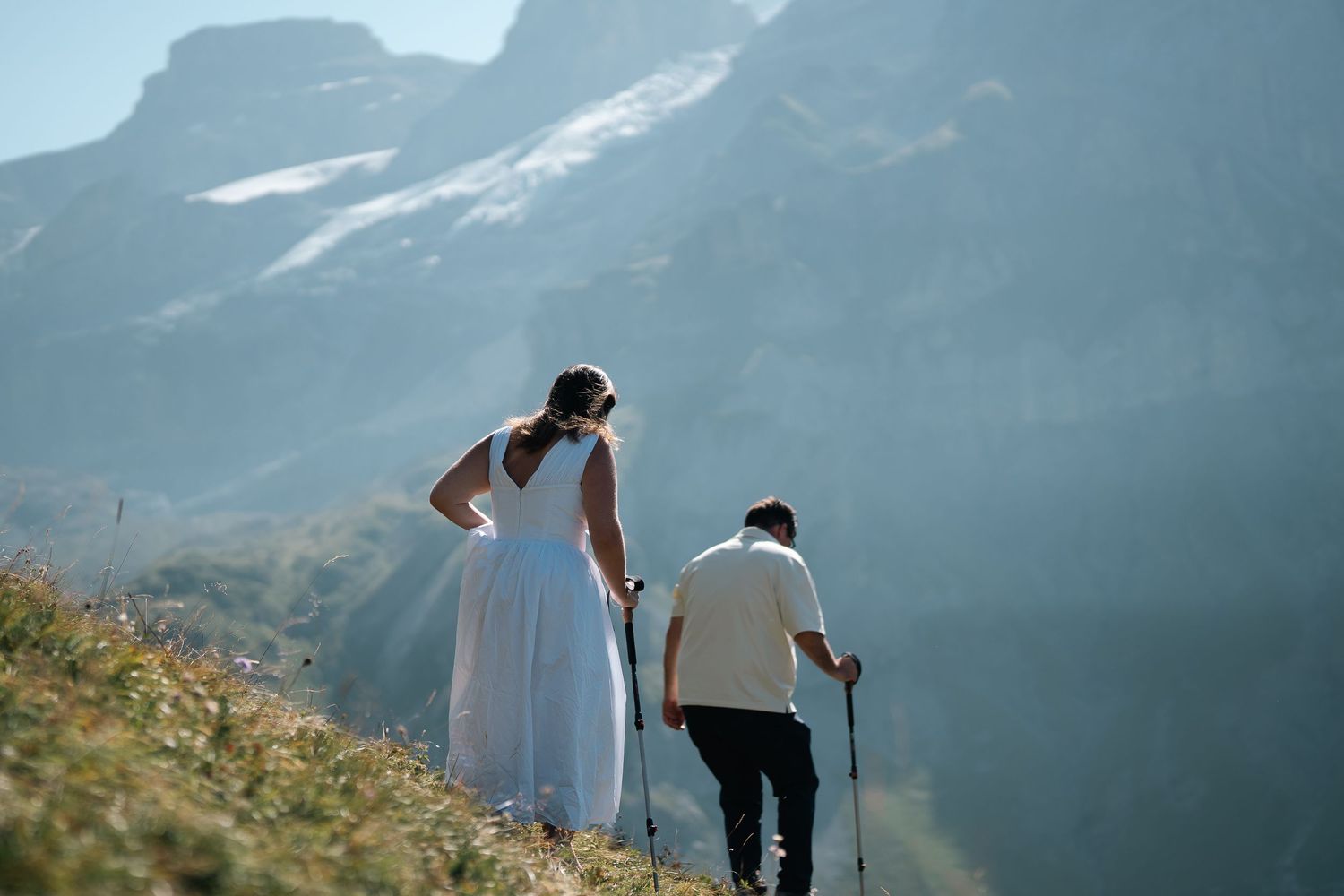 Bride in white gown and groom with hiking poles walk toward misty mountain peaks on a scenic alpine slope.