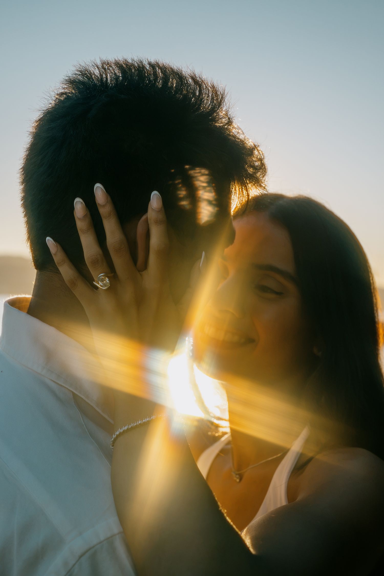 Couple sharing a romantic moment at golden hour, sun flare highlighting an engagement ring on her hand.