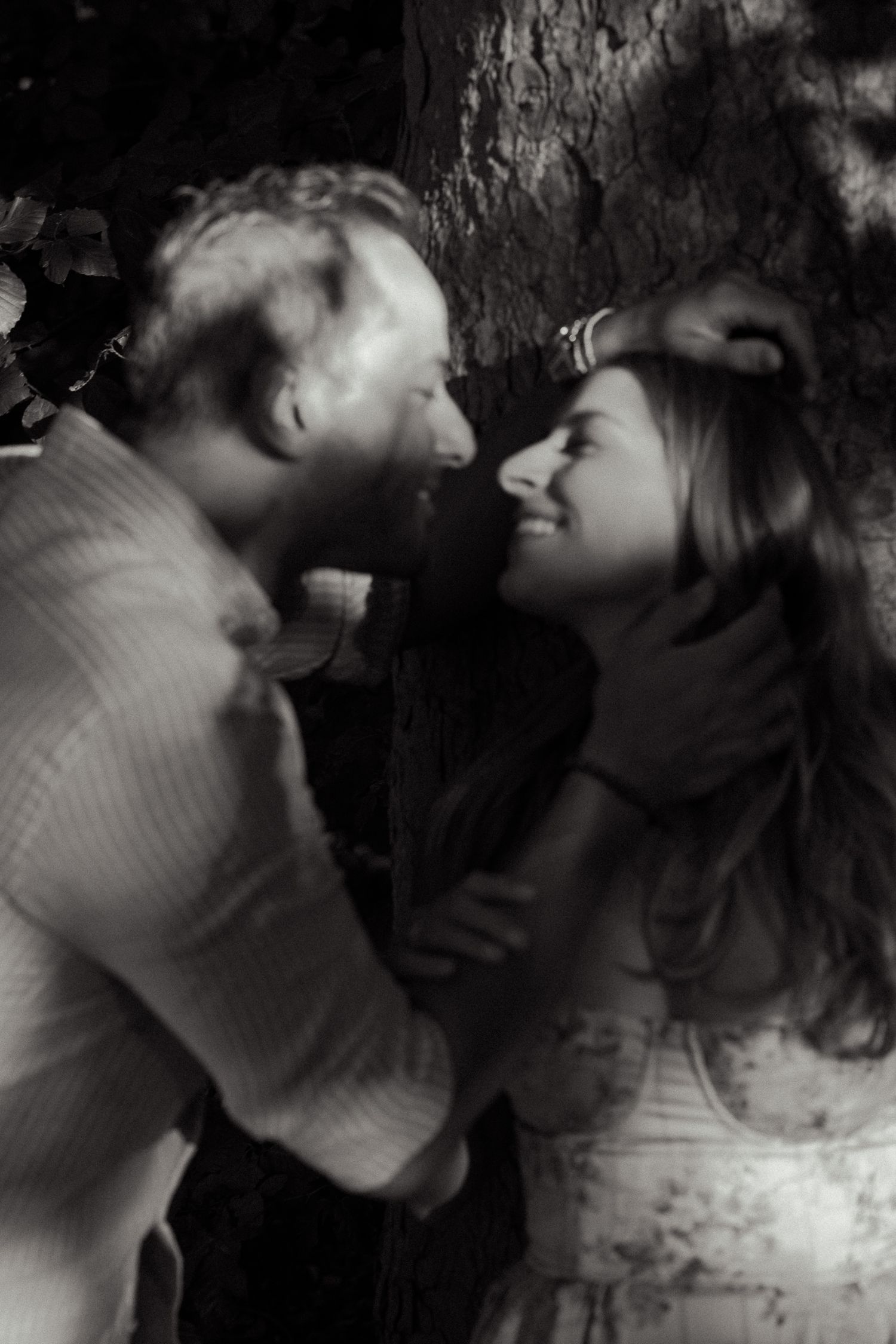 Black and white photo of a romantic couple smiling and gazing at each other against a tree backdrop.