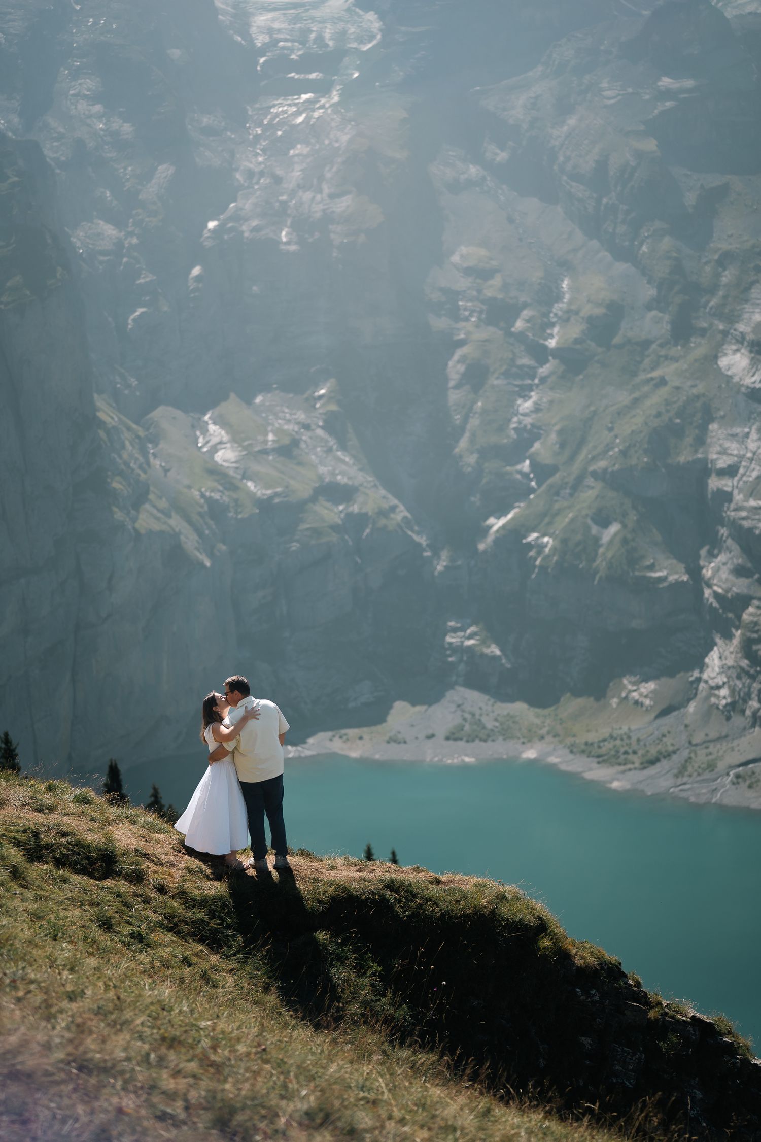 Couple embracing on a mountain cliff edge overlooking a turquoise alpine lake surrounded by rocky peaks.