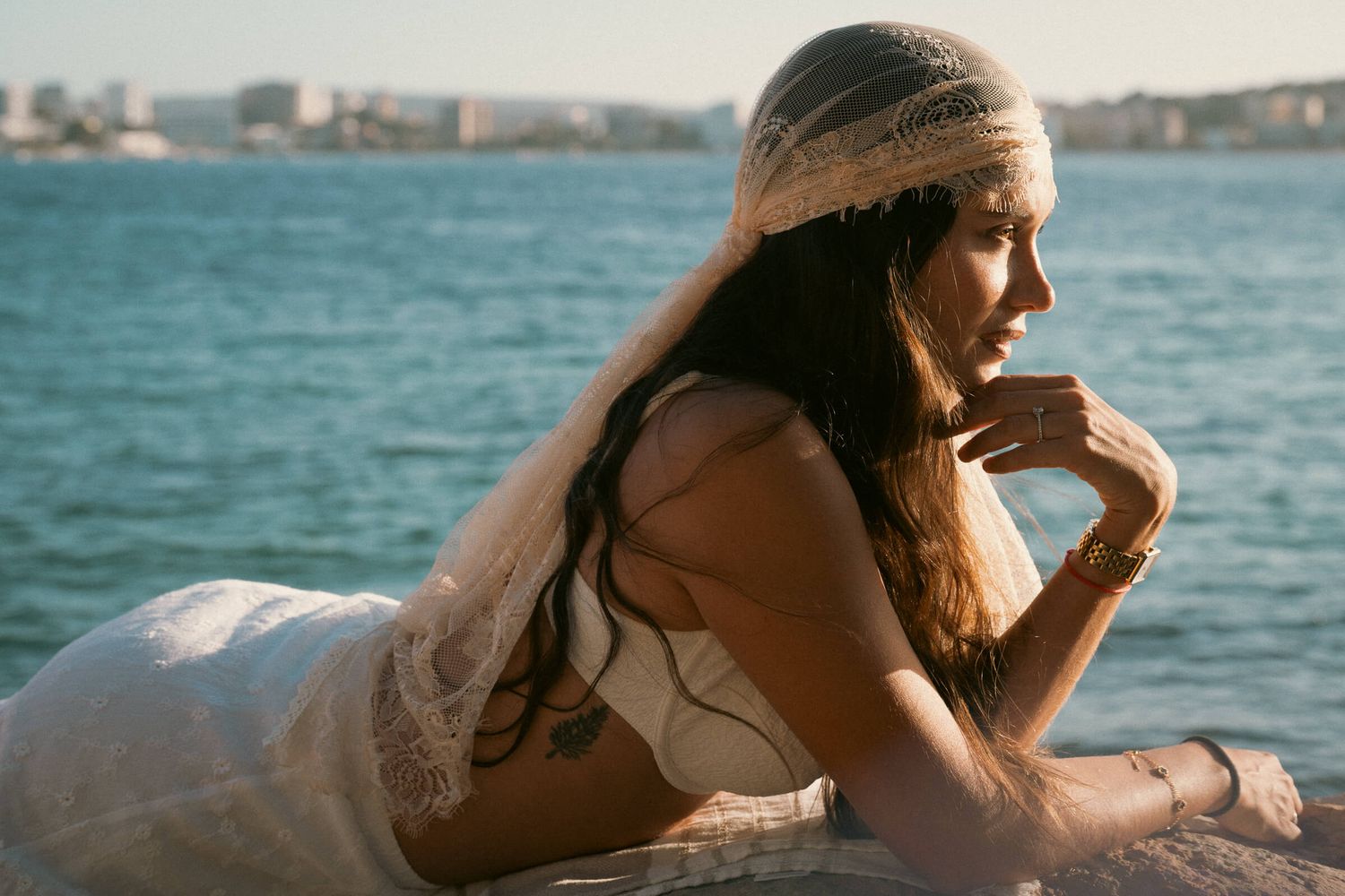 A woman in a white dress and lace headpiece gazes thoughtfully by the sea in warm golden light.