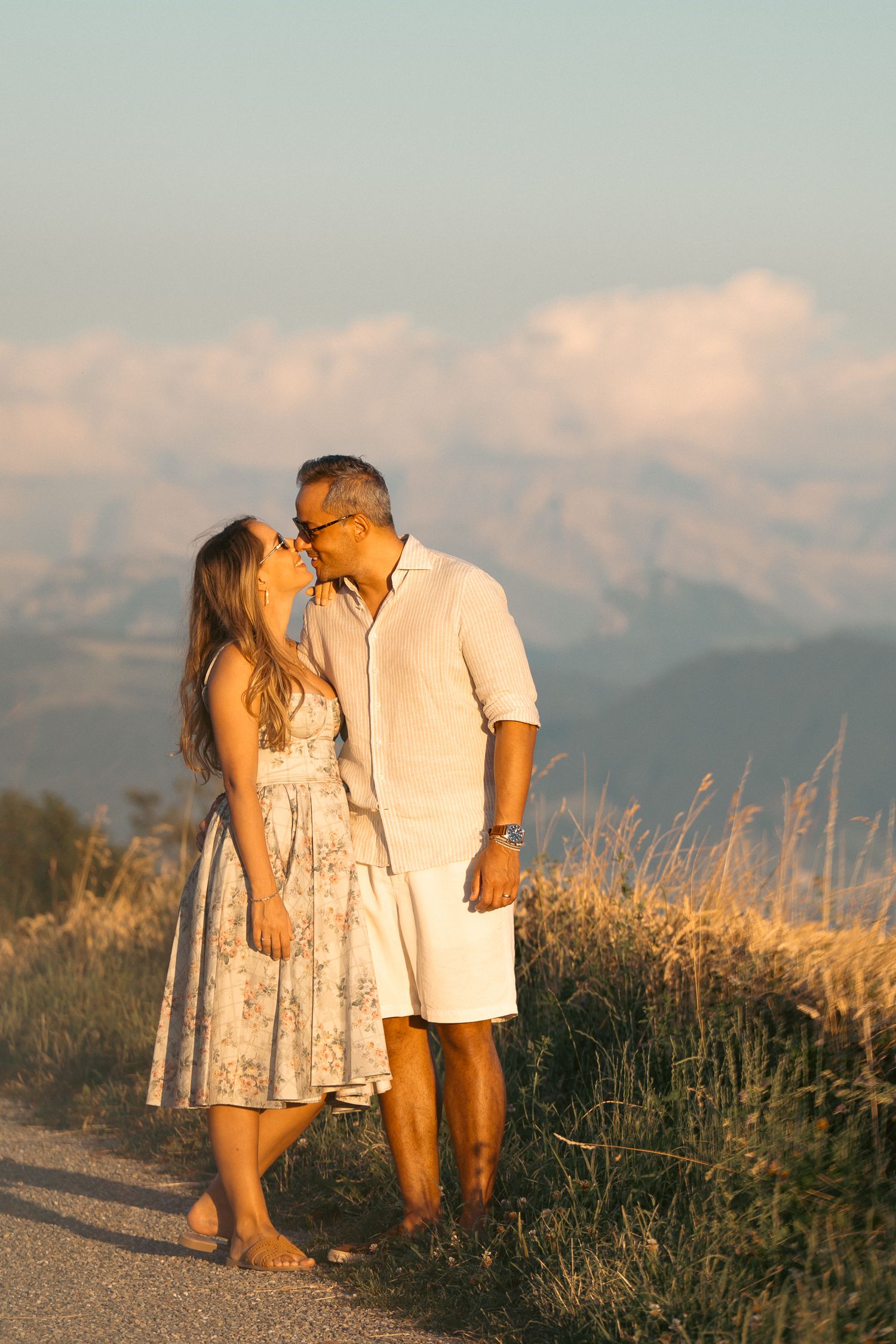 Romantic couple kissing on a scenic mountain path at golden hour, woman in floral dress, man in white.