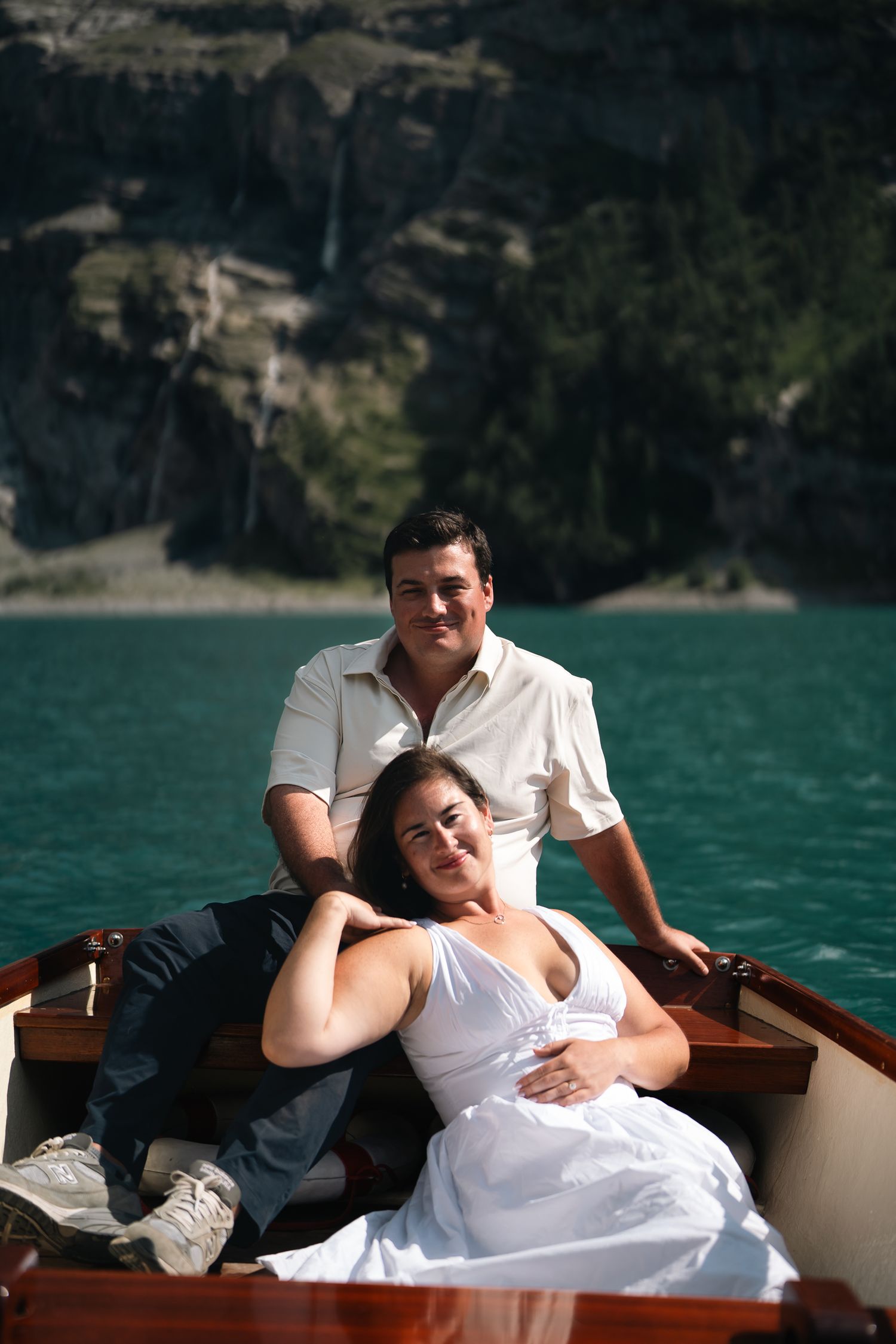 Couple relaxing in a wooden boat on turquoise water with rocky cliffs and a waterfall in the background.