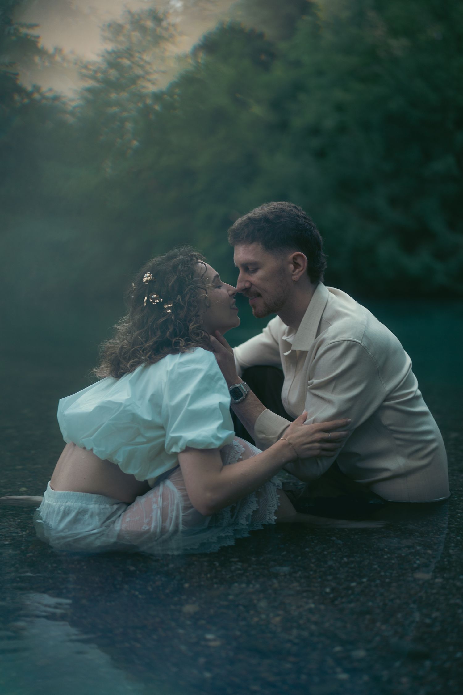 A couple sits intimately in shallow water, nearly kissing, surrounded by misty green trees in a romantic outdoor scene.