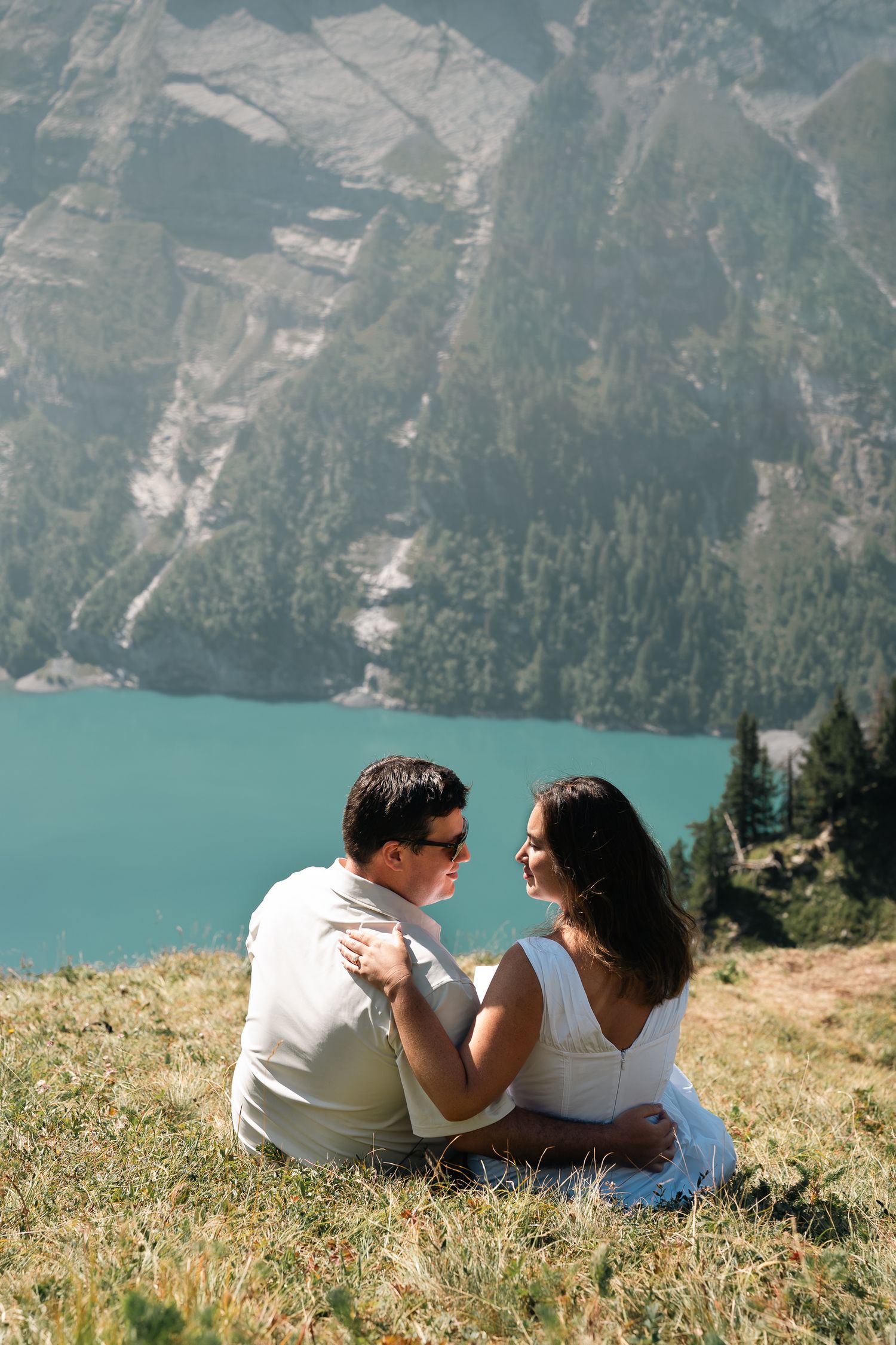 Couple sitting on a grassy hillside overlooking a turquoise alpine lake, sharing a romantic moment in the mountains.