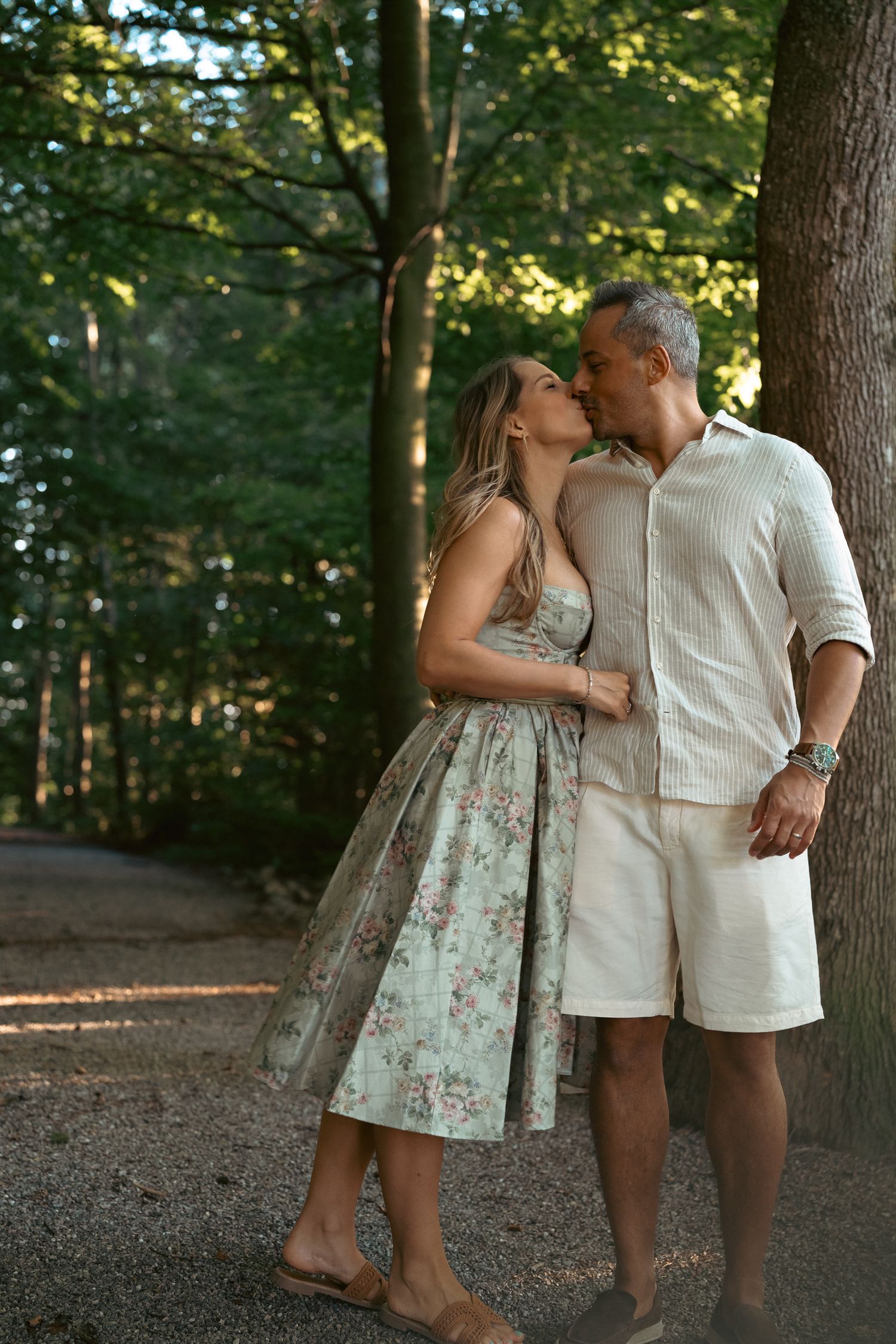 Romantic couple sharing a kiss beside a tree in a sunlit forest setting.
