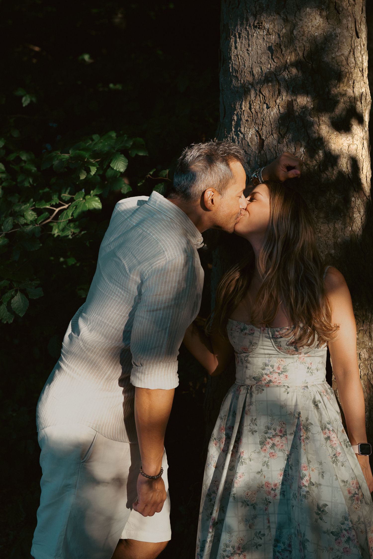 Couple sharing a romantic kiss near a tree in warm golden light, man in white shirt and woman in floral dress.