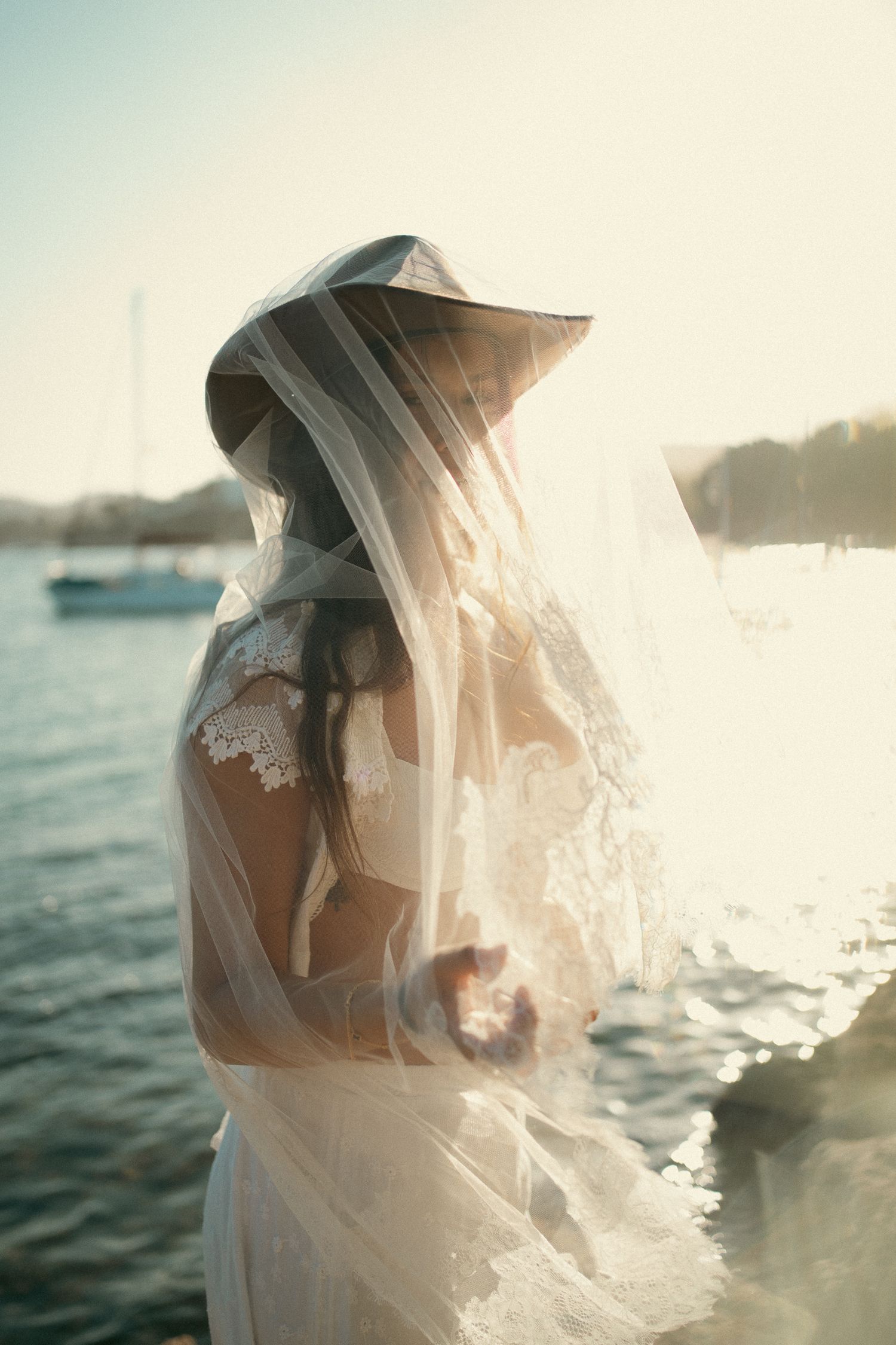 Bride in lace wedding dress and wide-brim hat with flowing veil by sunlit waterfront.
