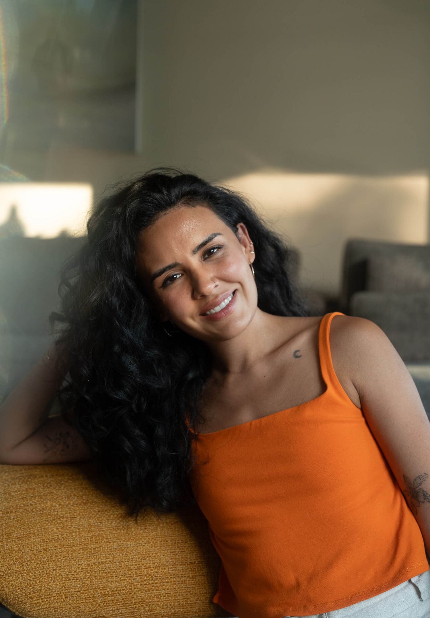 Smiling woman with curly dark hair wearing an orange tank top, relaxing on a sofa.