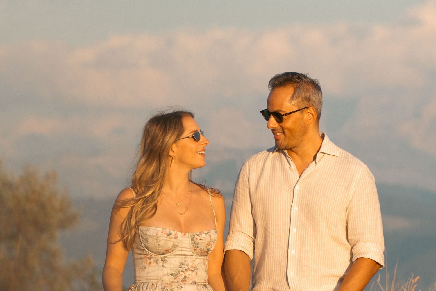 Couple smiling at each other outdoors at golden hour with mountains and clouds in the background.