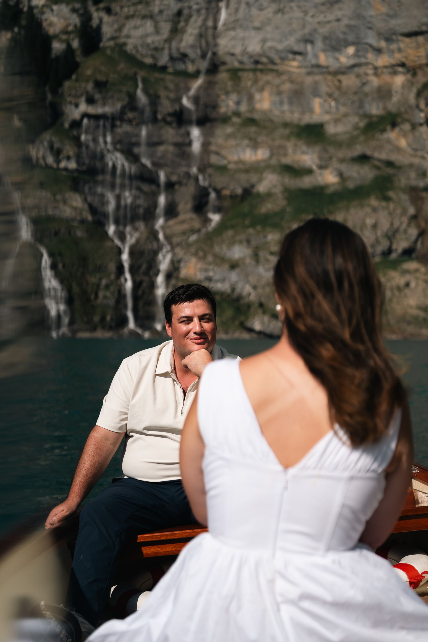 A smiling groom sits in a wooden boat facing his bride in a white backless dress.