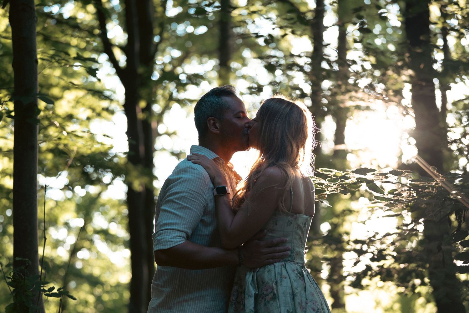 Couple sharing a romantic kiss in a sunlit forest, surrounded by lush green trees with golden light filtering through.