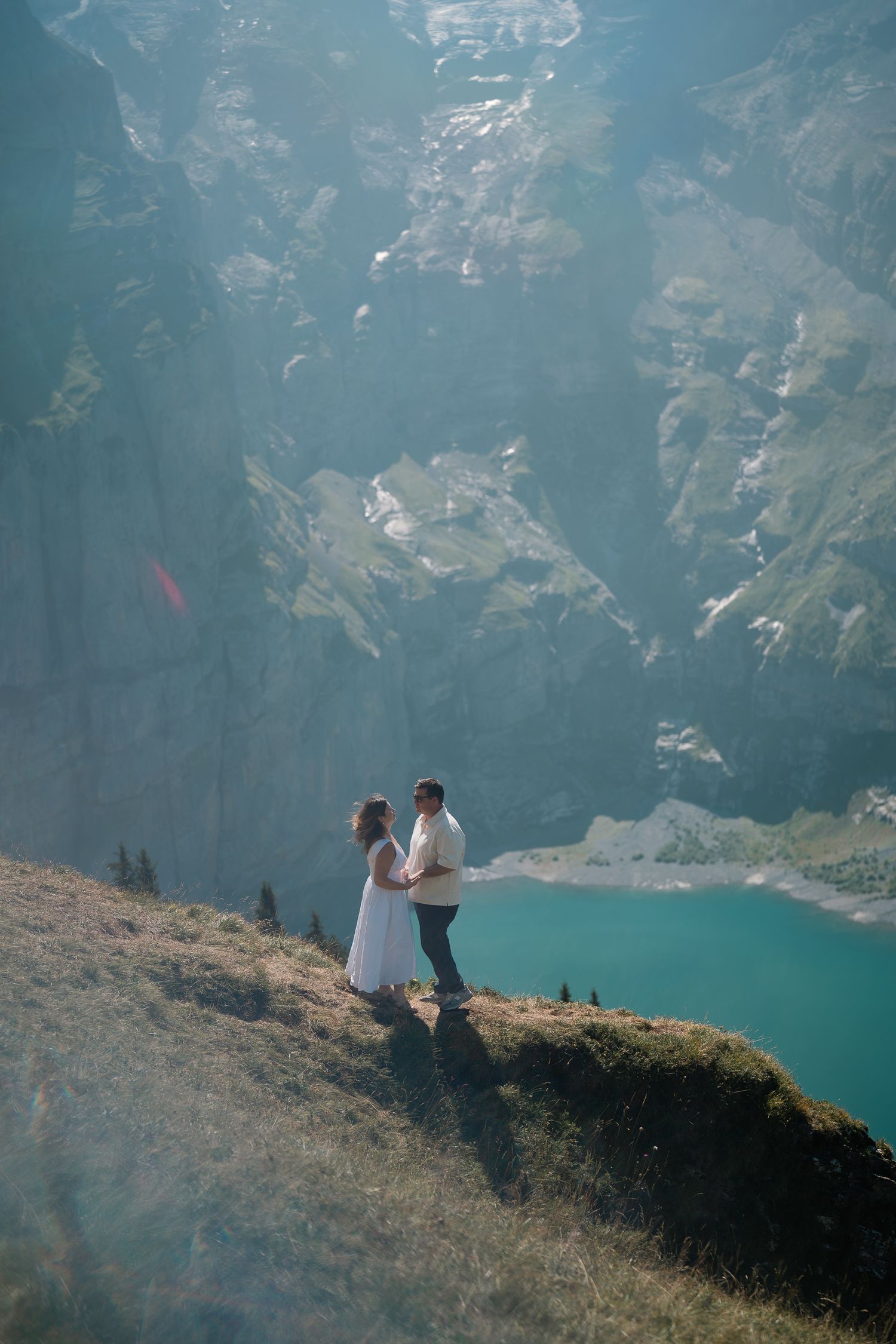 Couple standing on a mountain ridge above a turquoise alpine lake surrounded by dramatic rocky cliffs.