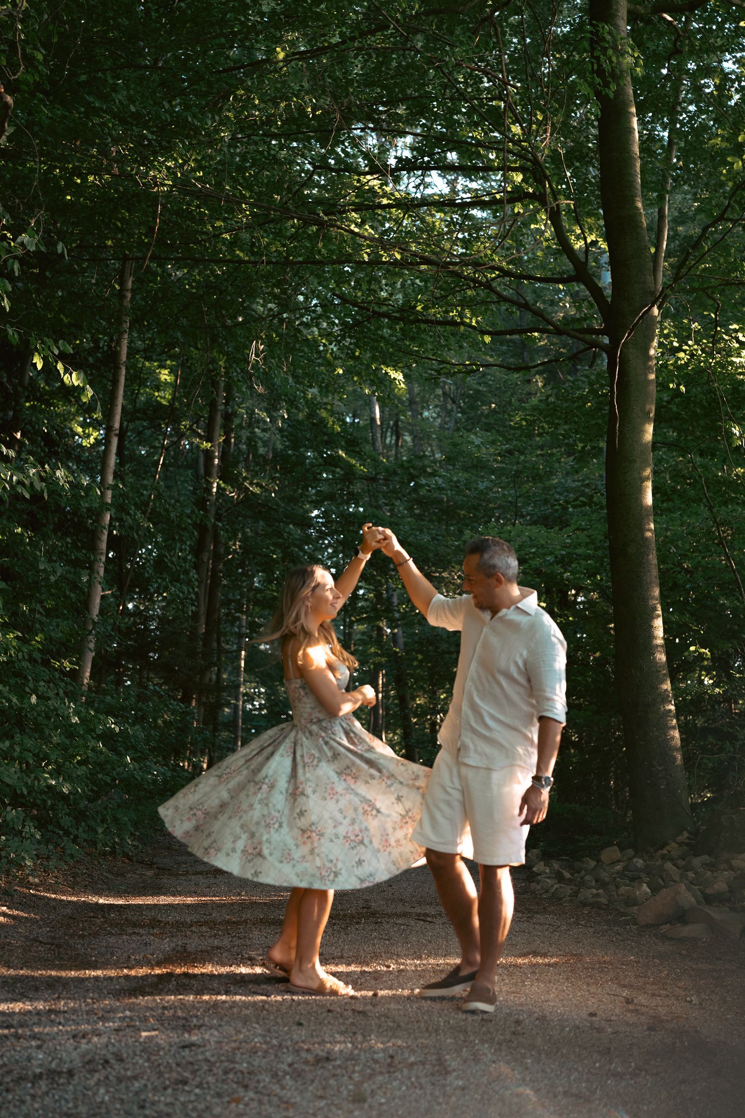 Couple dancing together on a sunlit forest path, woman in floral dress twirling.