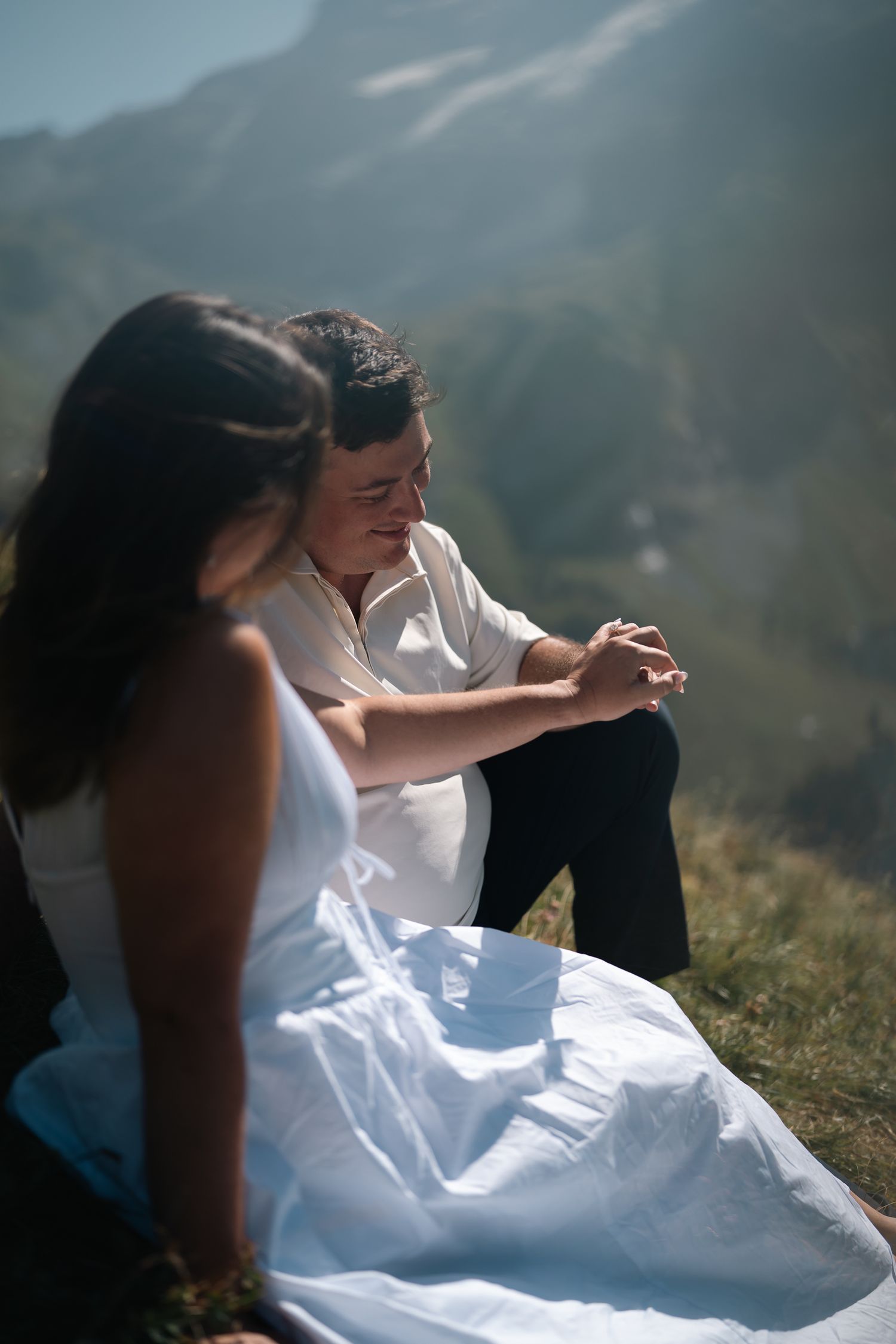 Couple in wedding attire sitting on a grassy hillside, bride in white dress, groom in white shirt and black pants.