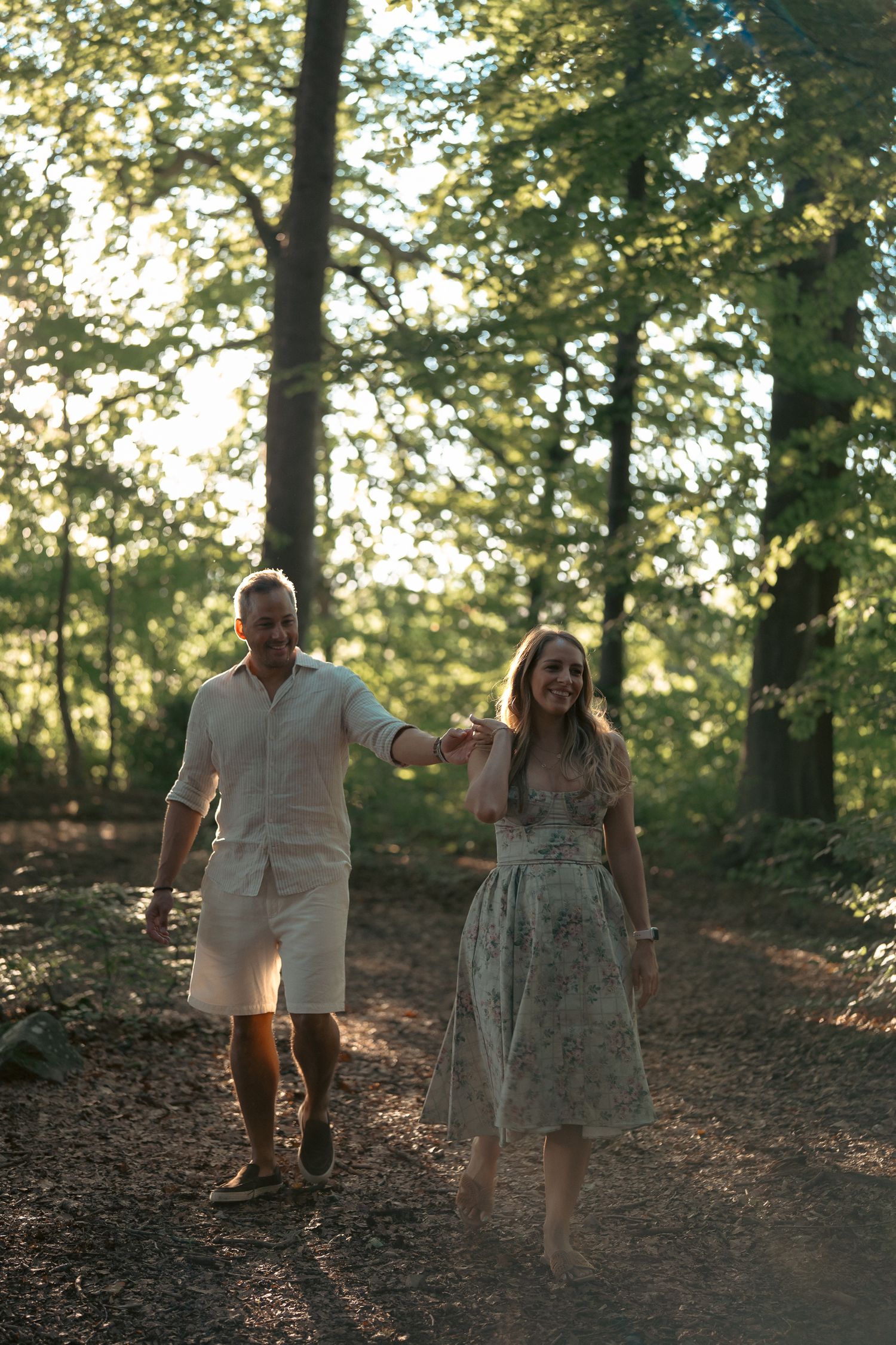 A couple holds hands walking on a sunlit forest path, smiling joyfully.