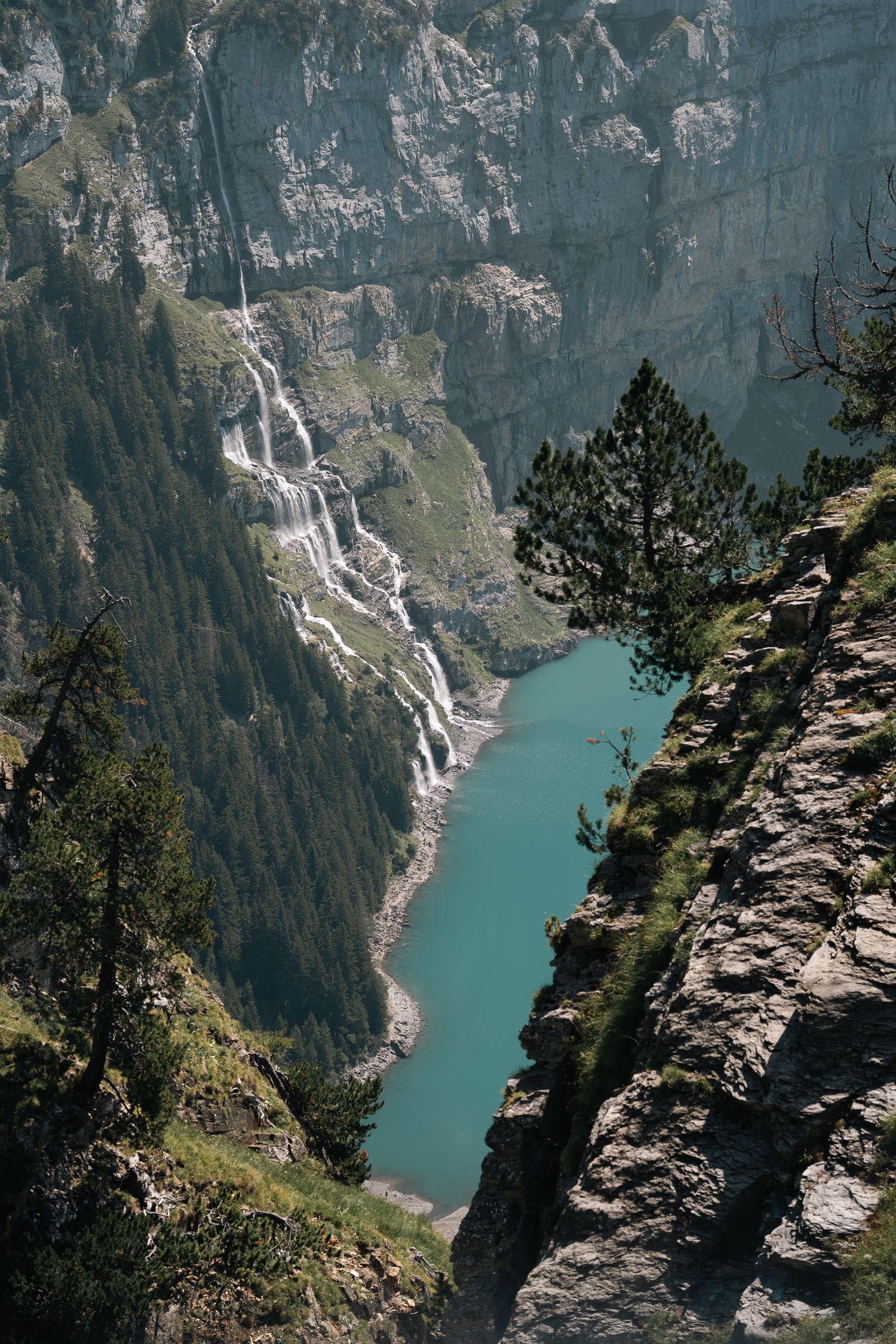 Stunning aerial view of turquoise alpine lake surrounded by rocky cliffs with cascading waterfalls and lush pine trees.
