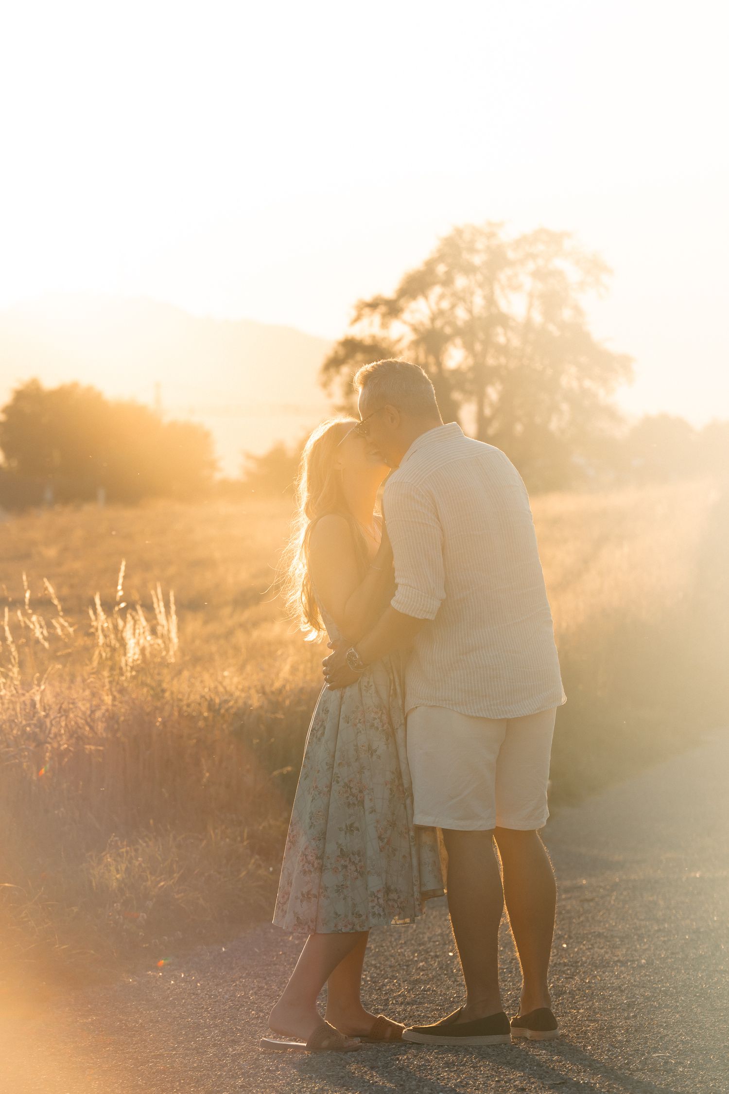 Couple sharing a tender moment on a sunlit country road surrounded by golden fields at sunset.