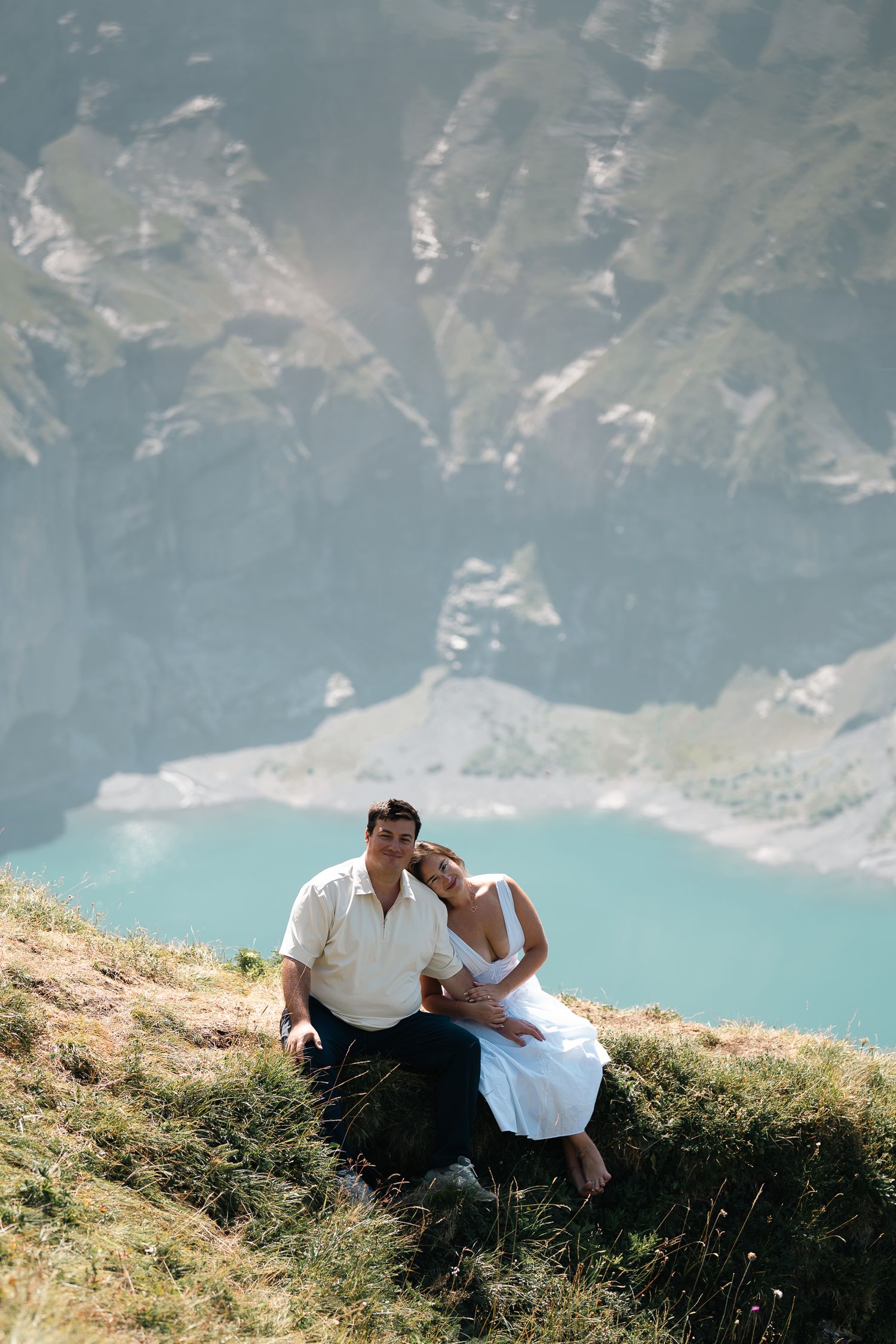 Couple sitting on a grassy mountain slope with a turquoise alpine lake and rocky peaks in the background.