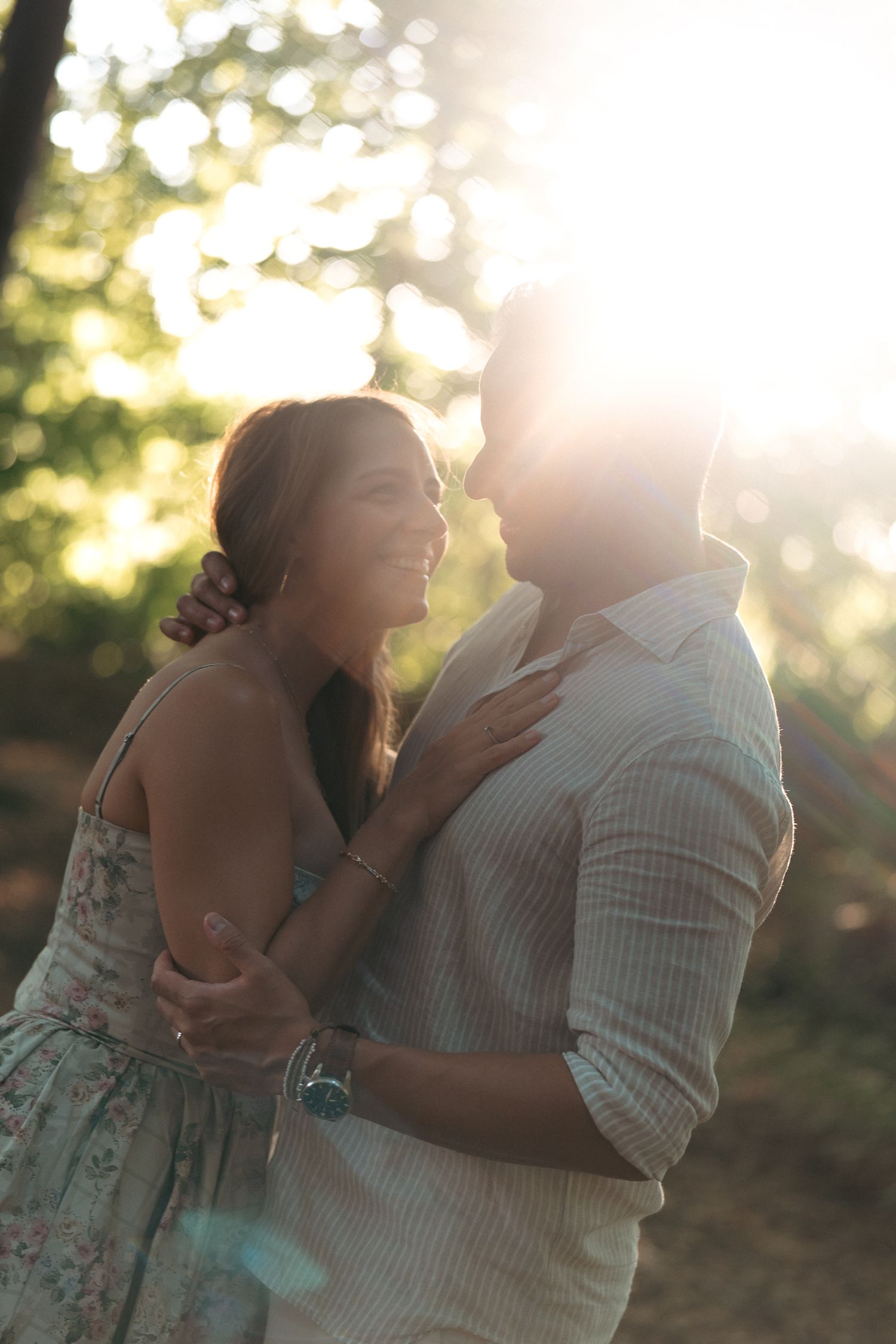 Couple embracing and smiling in golden sunlight among trees, woman in floral dress, romantic outdoor portrait.