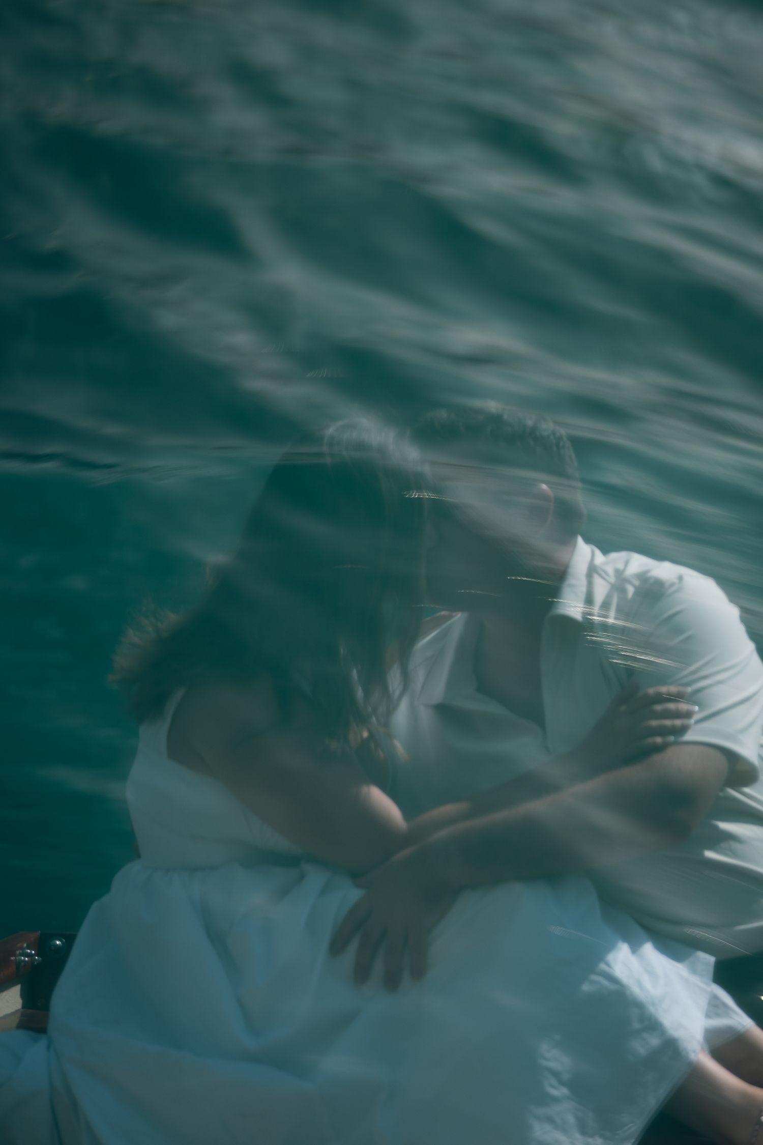 A couple in white clothing shares a tender embrace on a boat, reflected in the teal ocean water below.