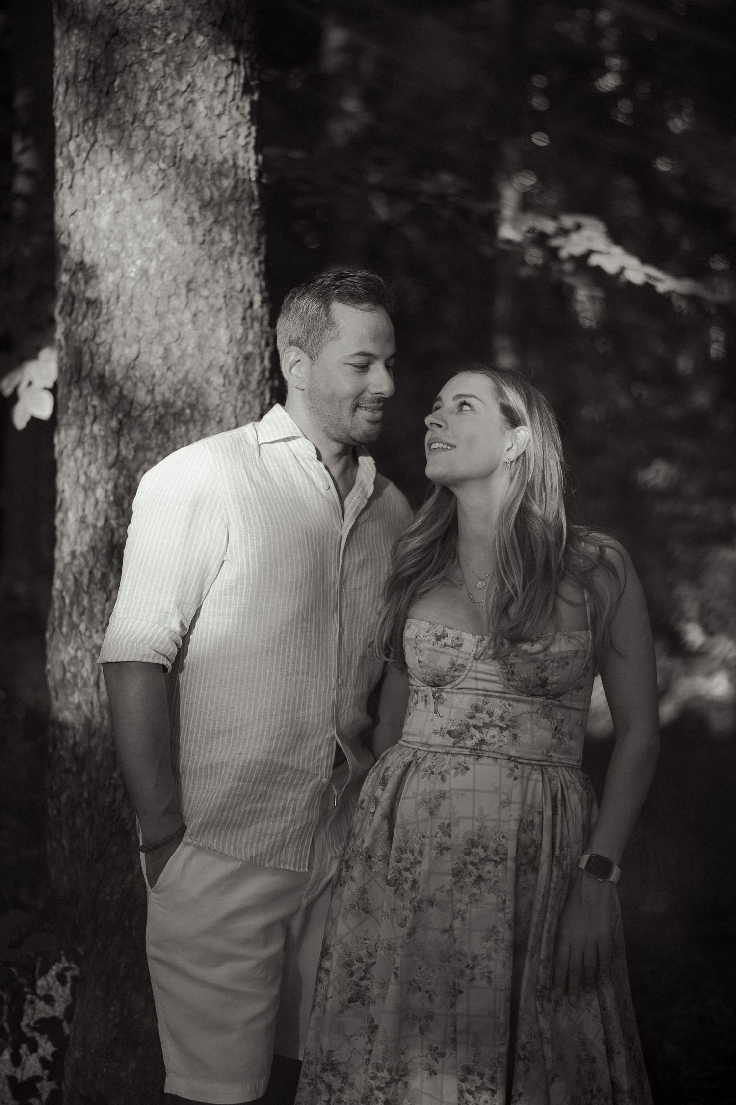 Black and white photo of a couple smiling at each other near a tree in a forest.