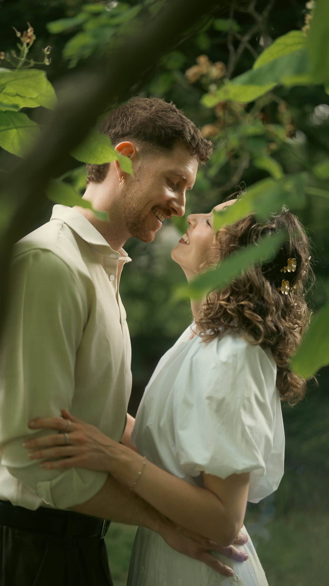A happy couple embraces and gazes at each other beneath green leafy branches in a garden.