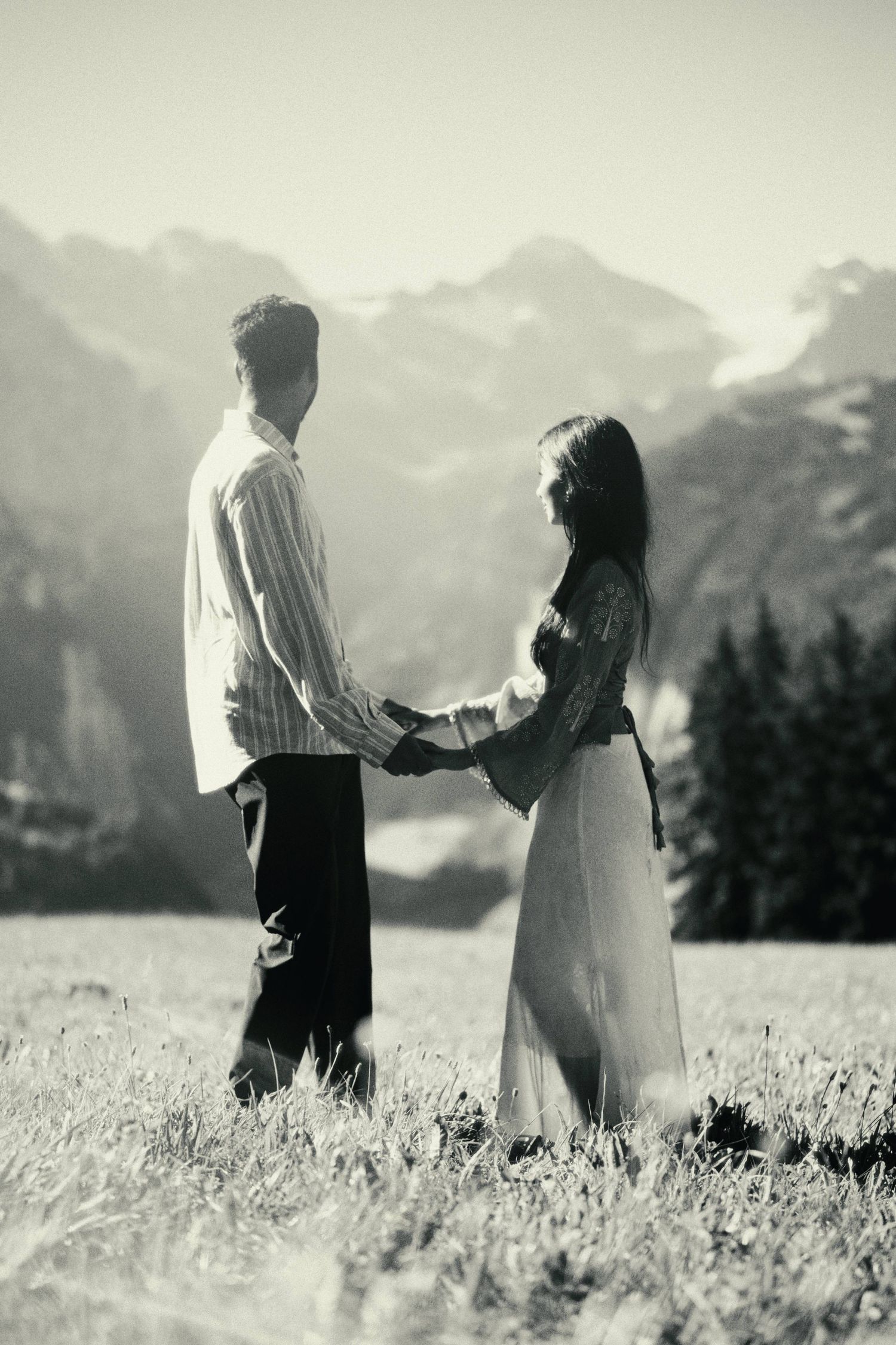 Black and white photo of a couple holding hands in a mountain meadow with snow-capped peaks in the background.