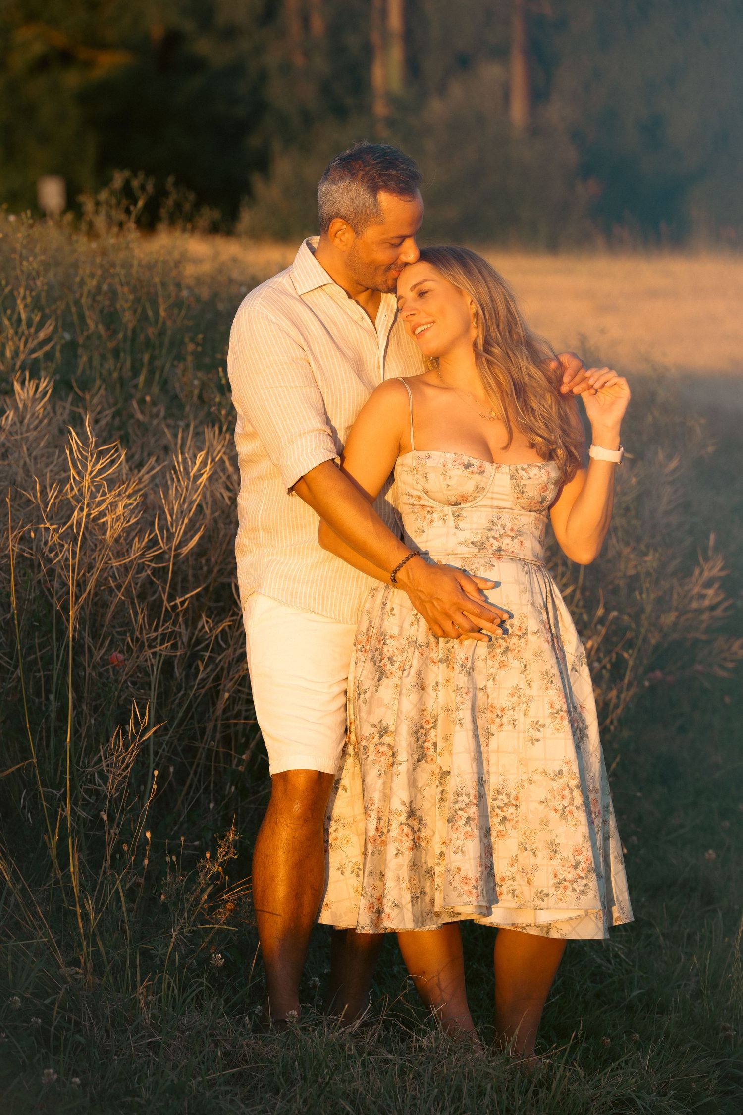 Romantic couple embracing in golden sunset light in a field, woman in floral dress smiling as man kisses her head.