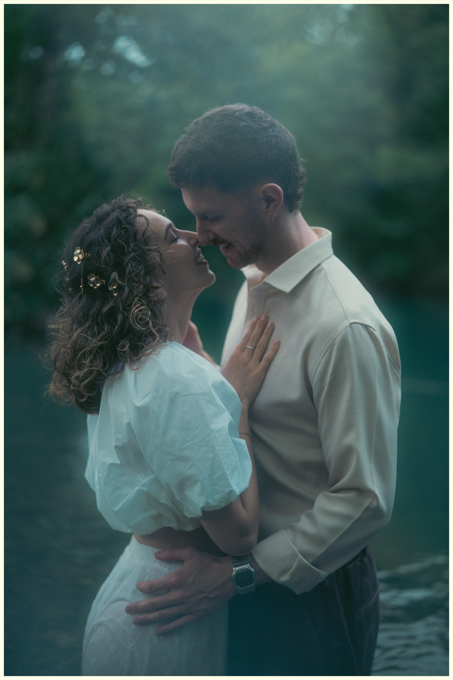 Couple sharing intimate moment near misty blue lake, bride in white dress touching grooms chest.