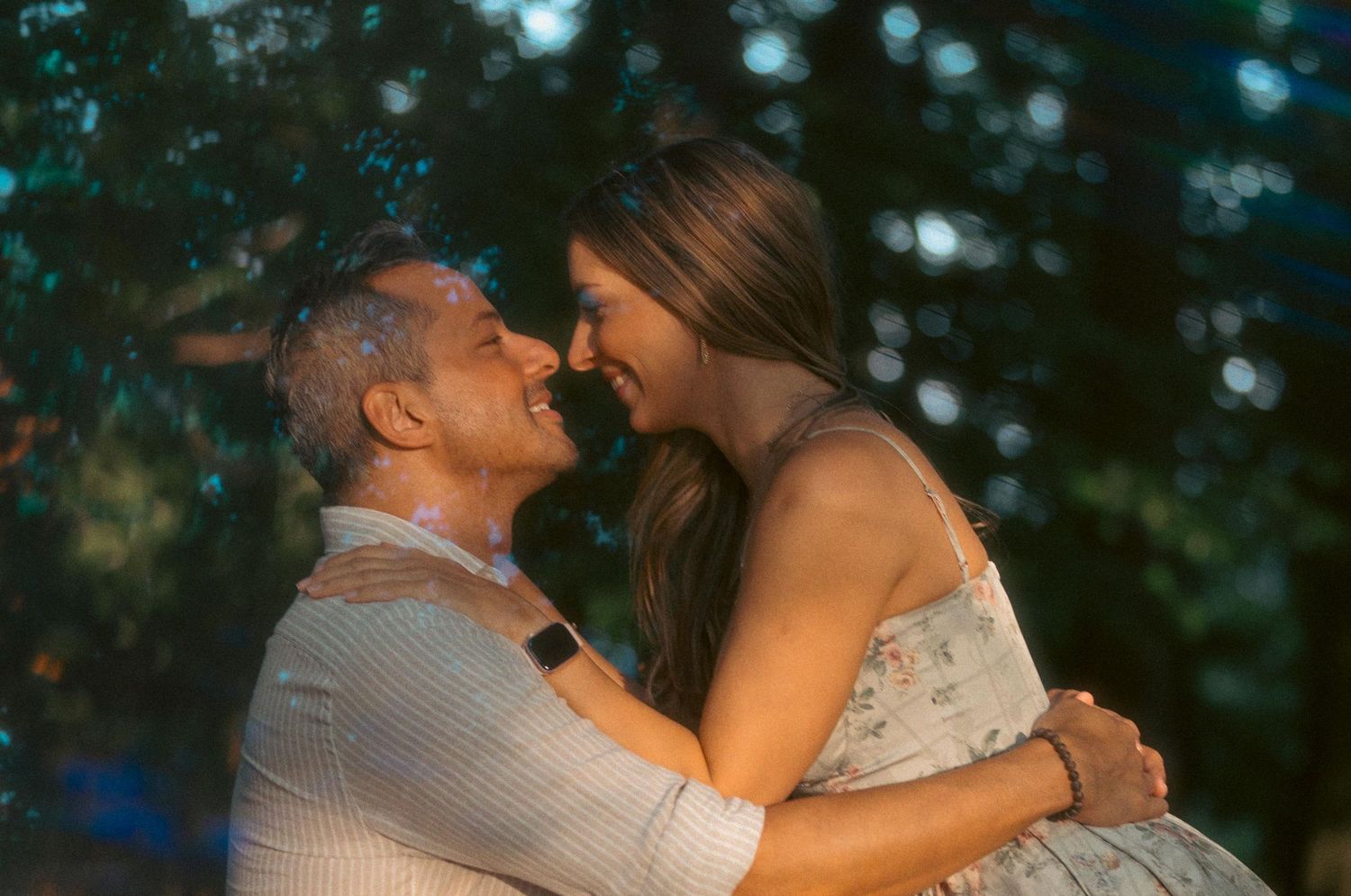 A couple sharing a romantic moment, smiling and embracing outdoors with lush green trees in the background.