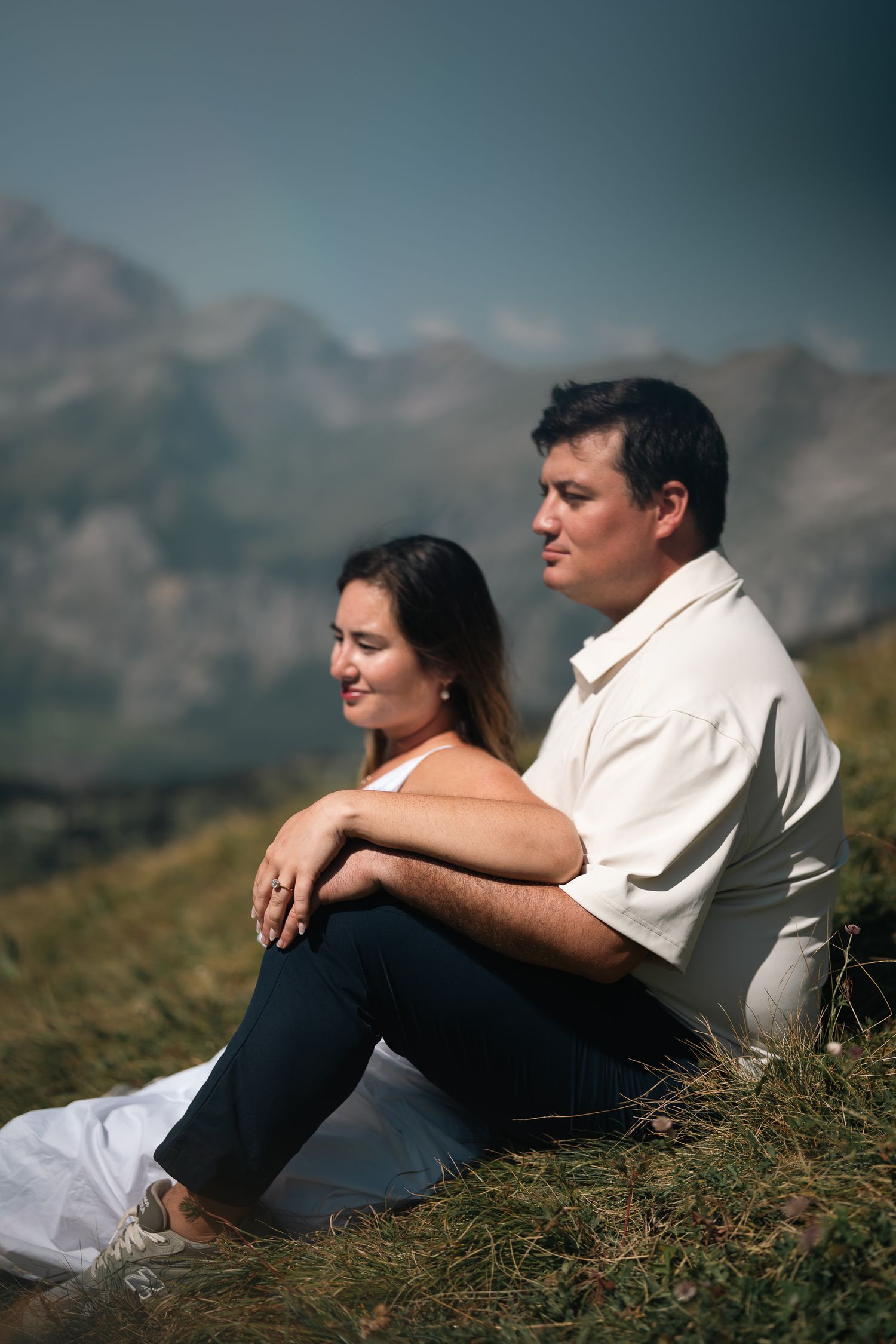 Couple sitting together on a grassy mountain slope, relaxing with scenic mountain views behind them.