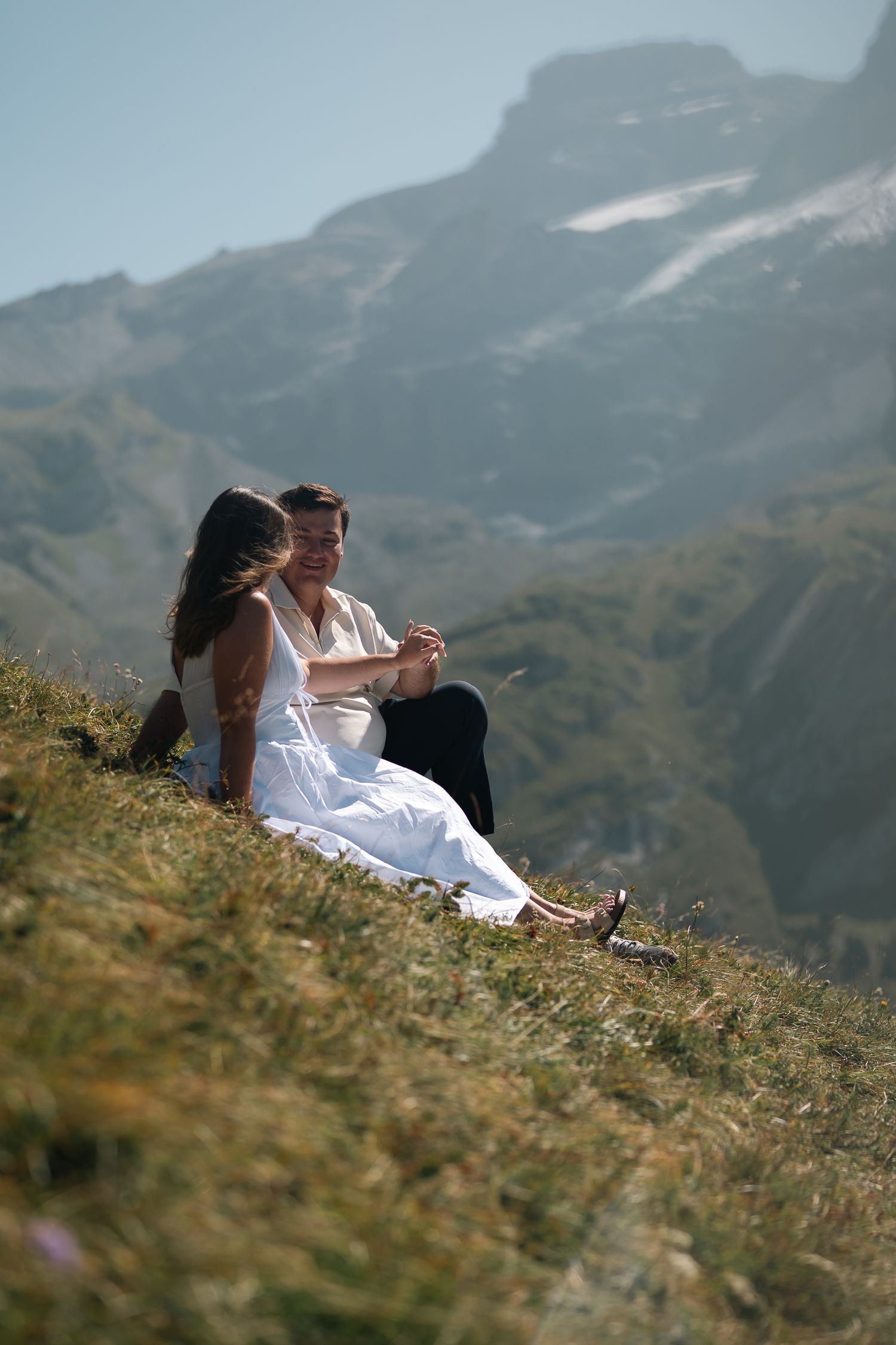 Couple sitting together on a grassy alpine hillside with snow-capped mountains in the background on a sunny day.