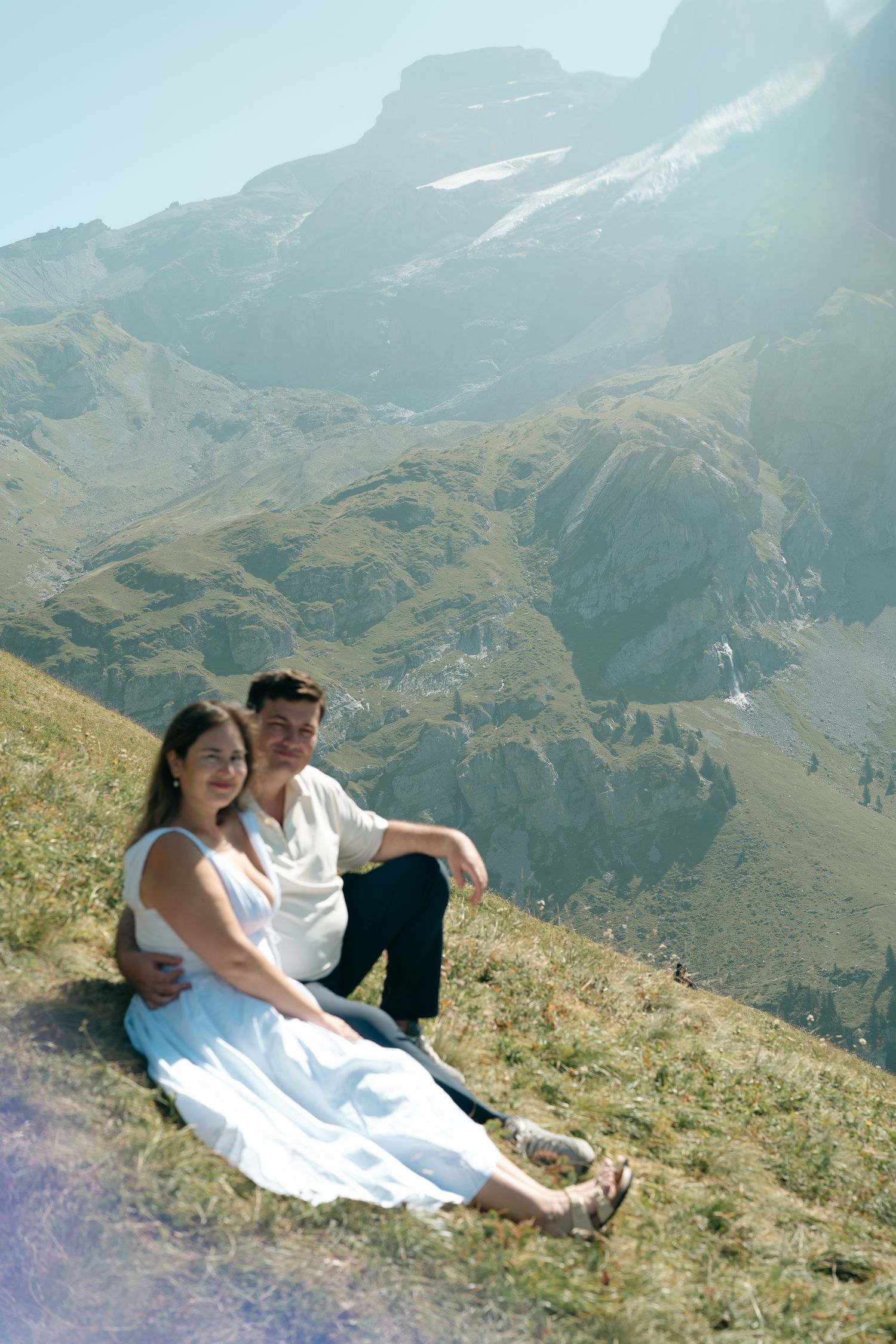 Couple sitting on alpine meadow with majestic mountain peaks in background on sunny day.