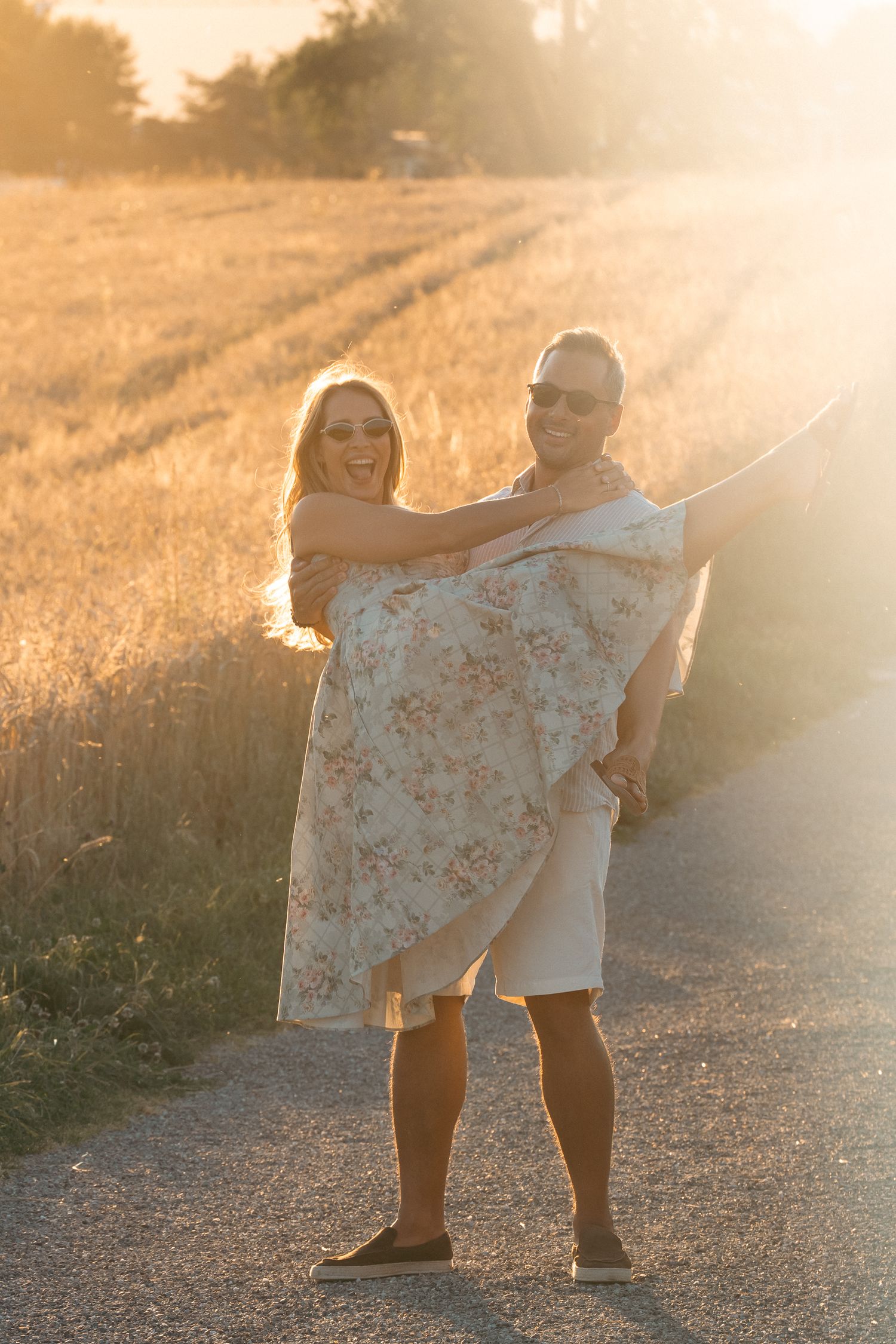 Couple laughing in golden sunset light on rural road, man carrying woman in floral dress near wheat field.