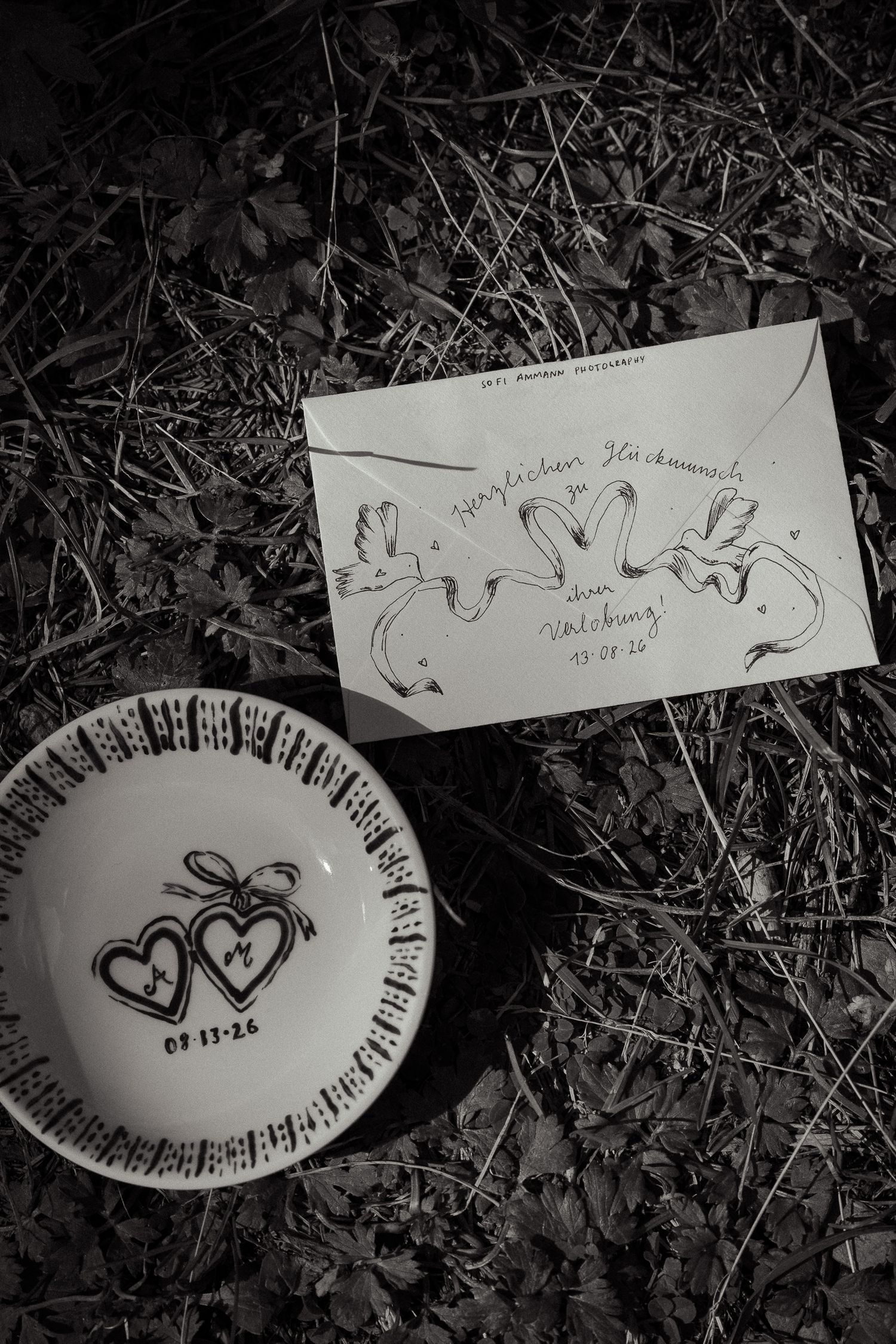 Black and white photo of a decorative wedding plate and envelope with swan illustrations on grass.
