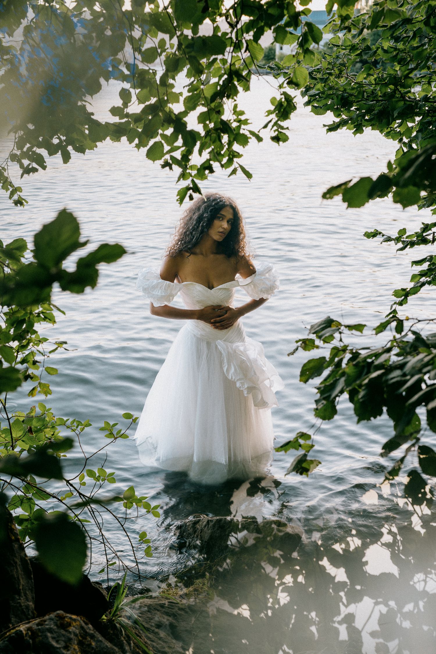 Woman in white off-shoulder wedding gown standing in a serene lake surrounded by lush green trees.