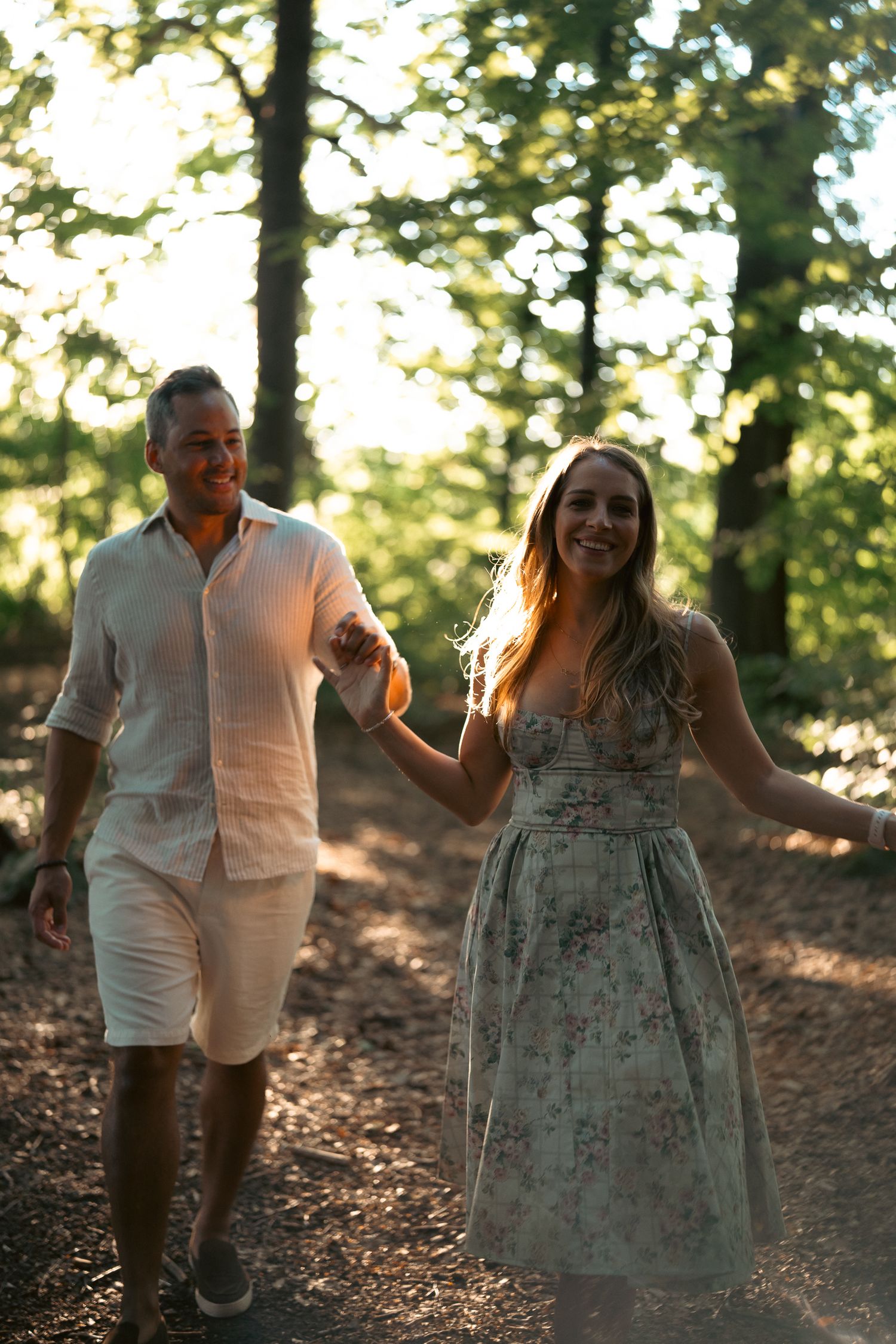A smiling couple holds hands while walking on a sunlit forest trail, the woman in a floral dress and man in white linen.