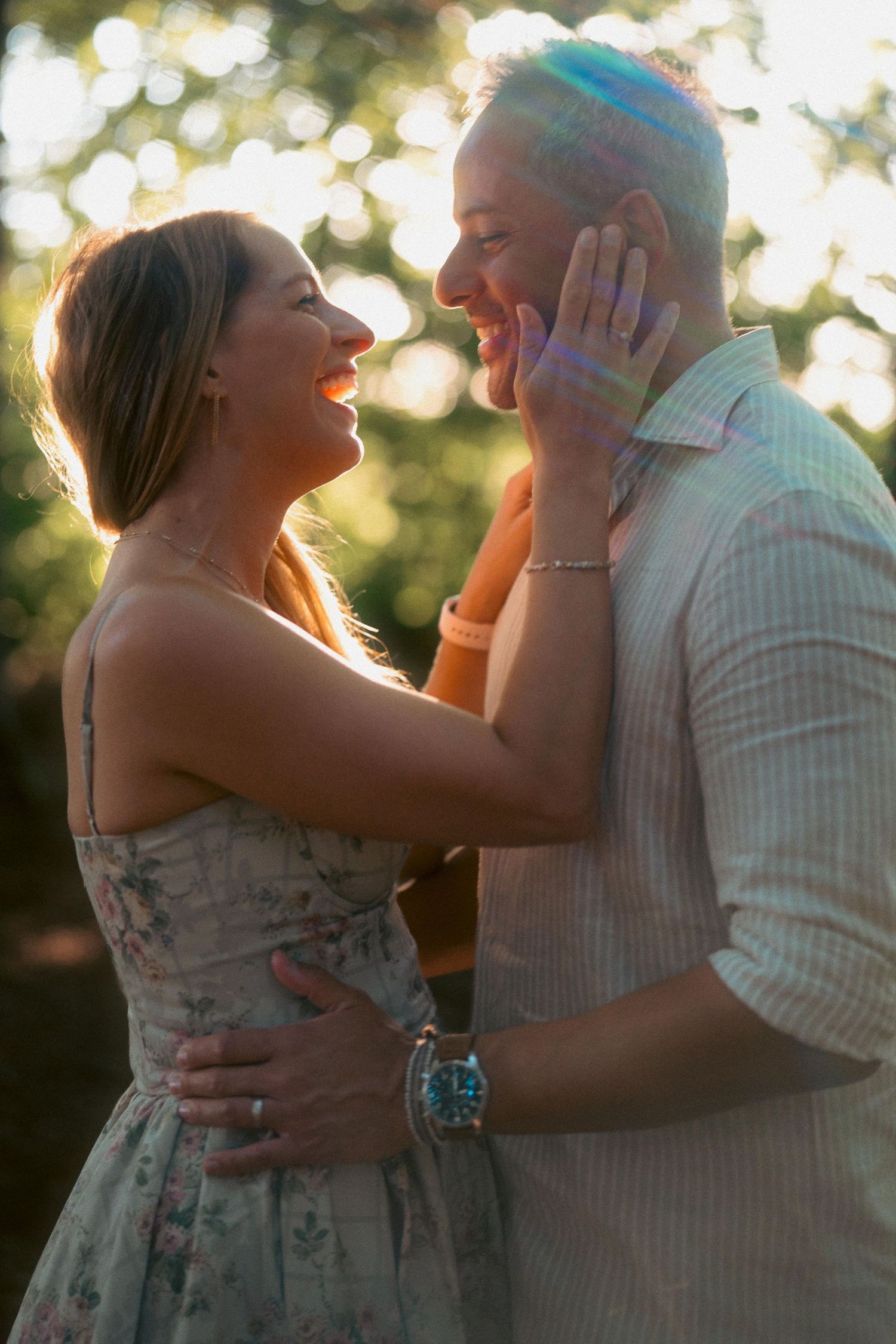 Happy couple embracing and laughing at sunset, woman in floral dress touching mans face tenderly.