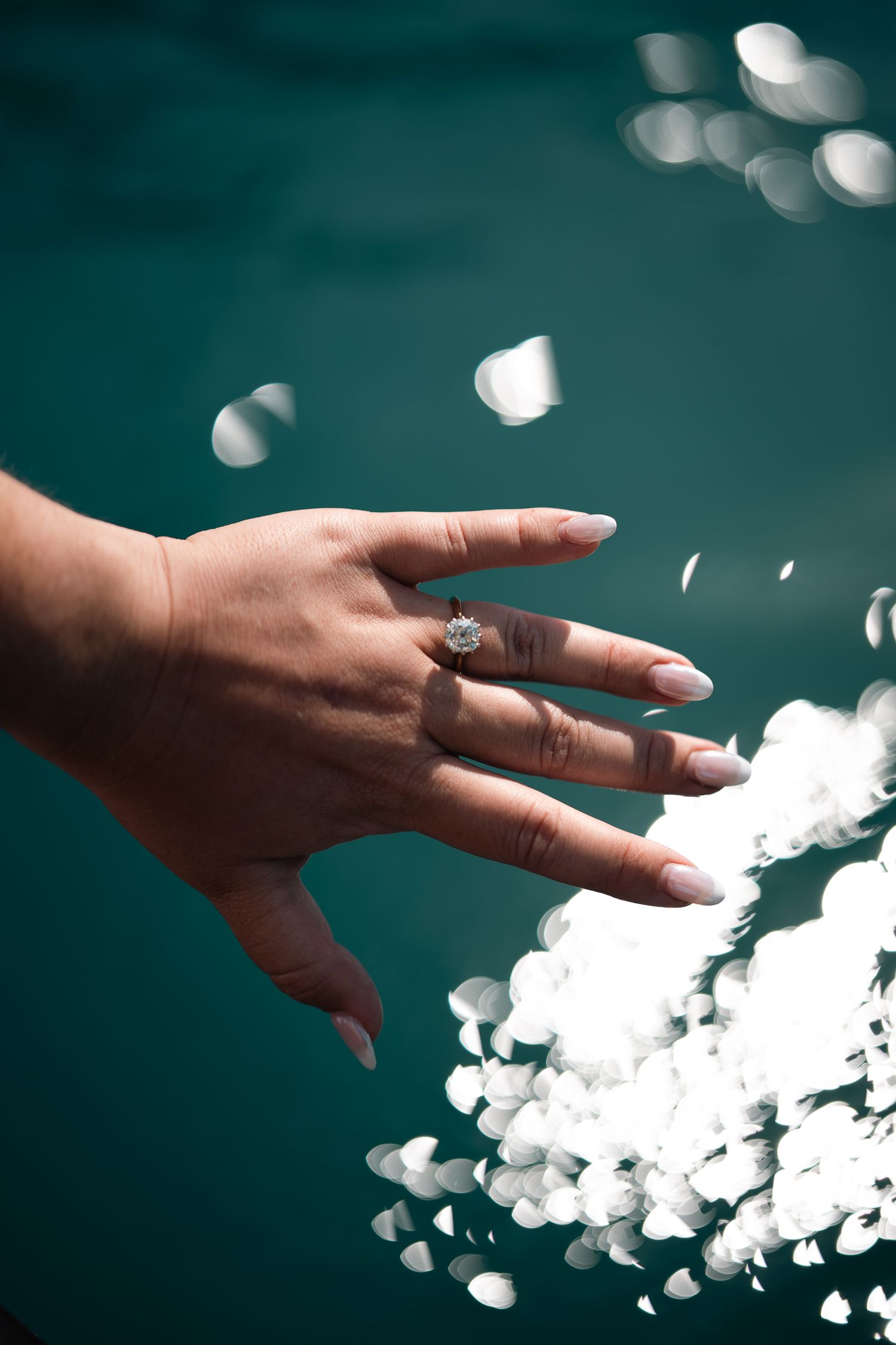 A hand with manicured nails wearing a diamond engagement ring, surrounded by floating white petals on teal background.