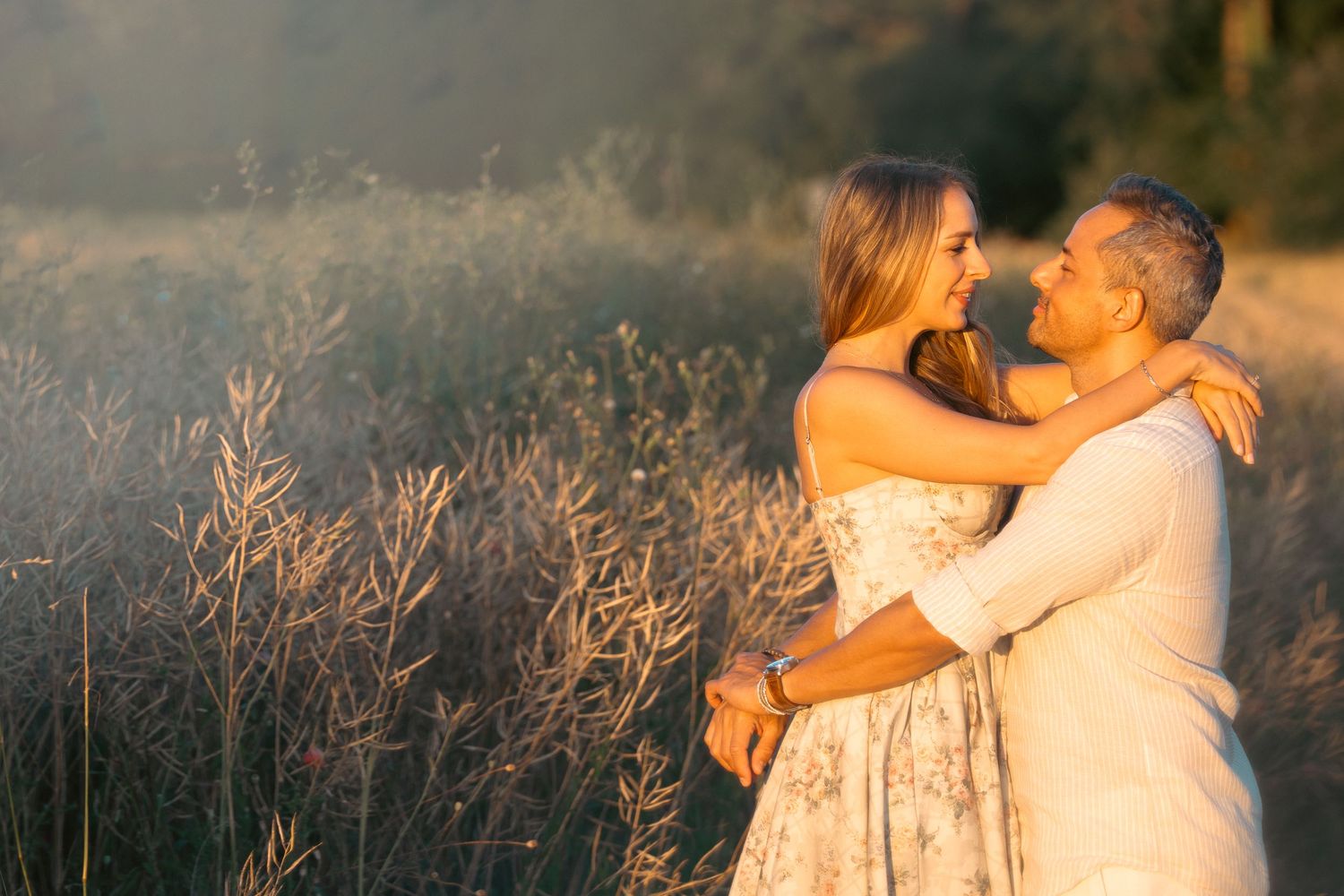 Happy couple embracing and smiling in a sunlit field of tall golden grasses during sunset.