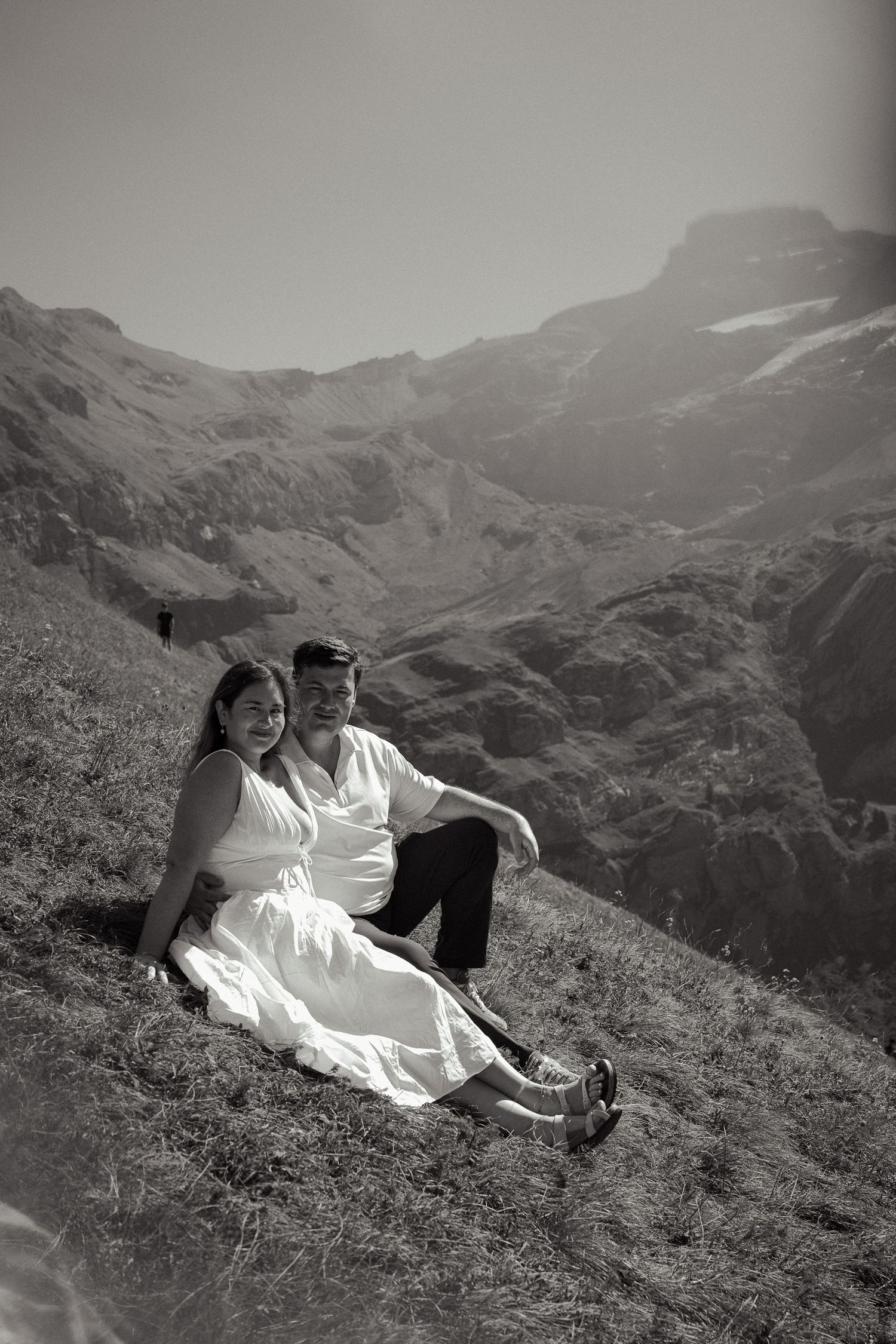 Black and white photo of a couple sitting on a grassy mountain slope, woman in white dress, scenic alpine backdrop.