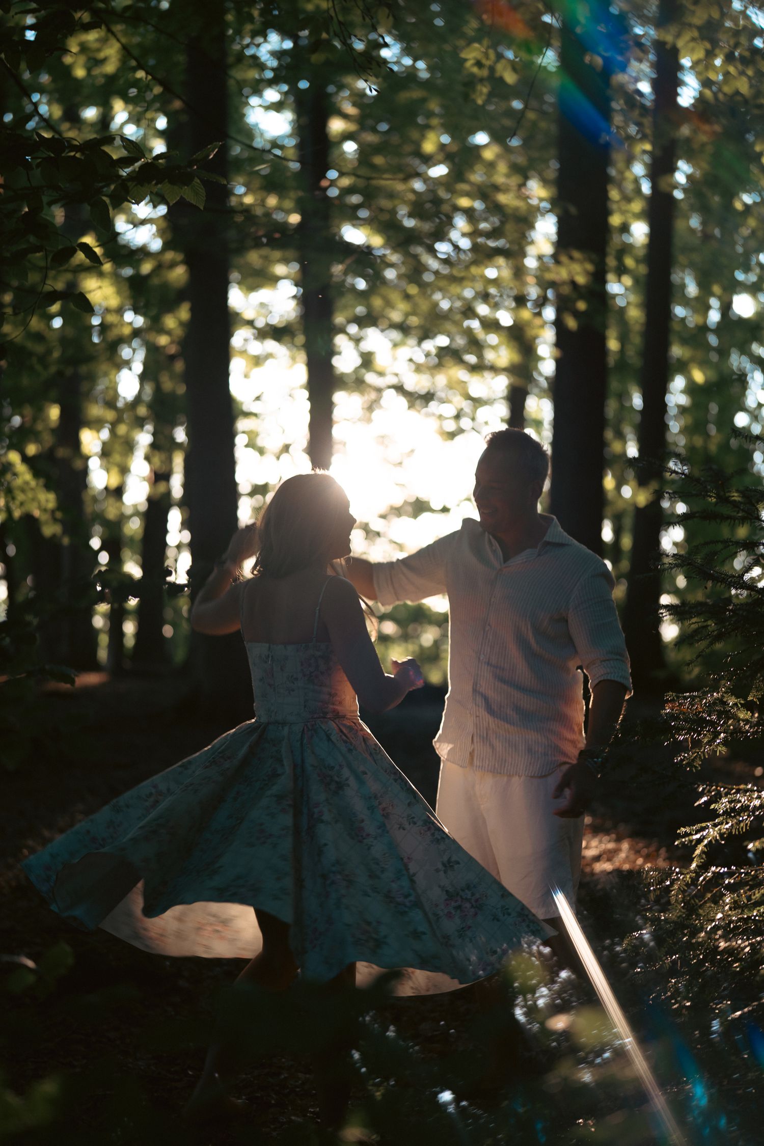 Couple dancing in a sunlit forest, woman in floral dress spinning gracefully among tall trees.