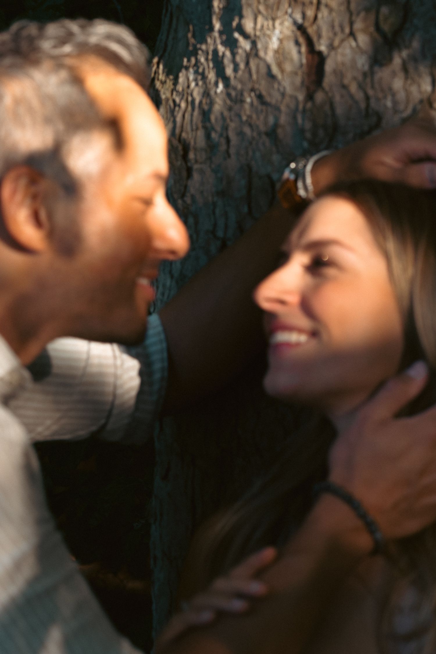 Couple smiling at each other tenderly against a tree trunk in soft warm light during golden hour.