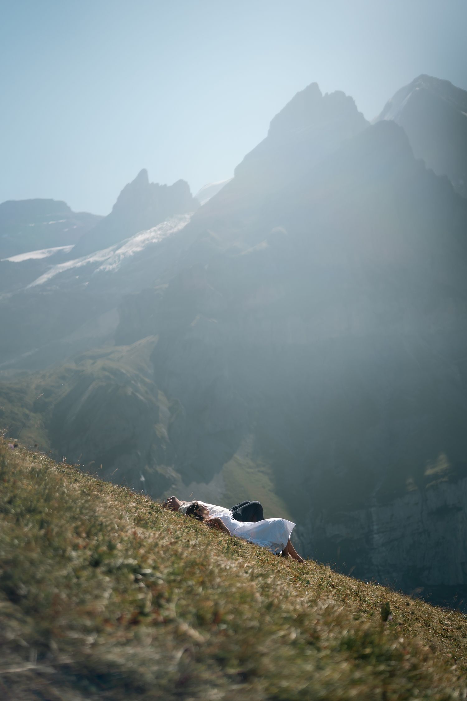 Person lying on a grassy mountain slope with dramatic rocky peaks and misty sunlight in the background.