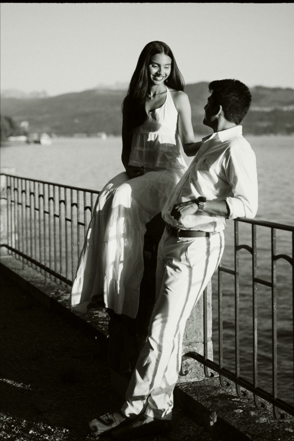 Black and white photo of a couple in white outfits sharing a romantic moment by a lakeside railing.