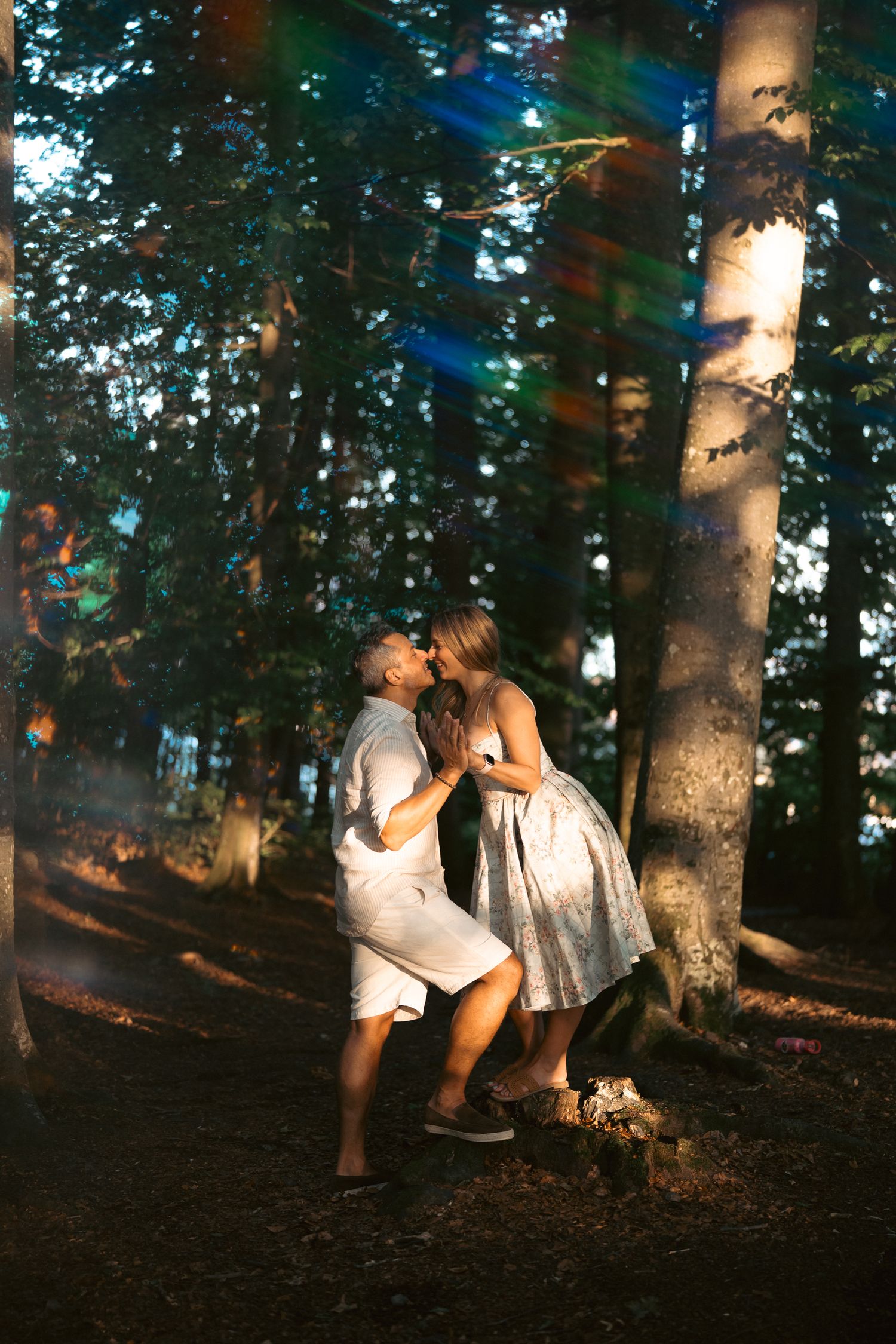 A couple shares a romantic kiss among tall forest trees with colorful light streaks in a dreamy outdoor setting.