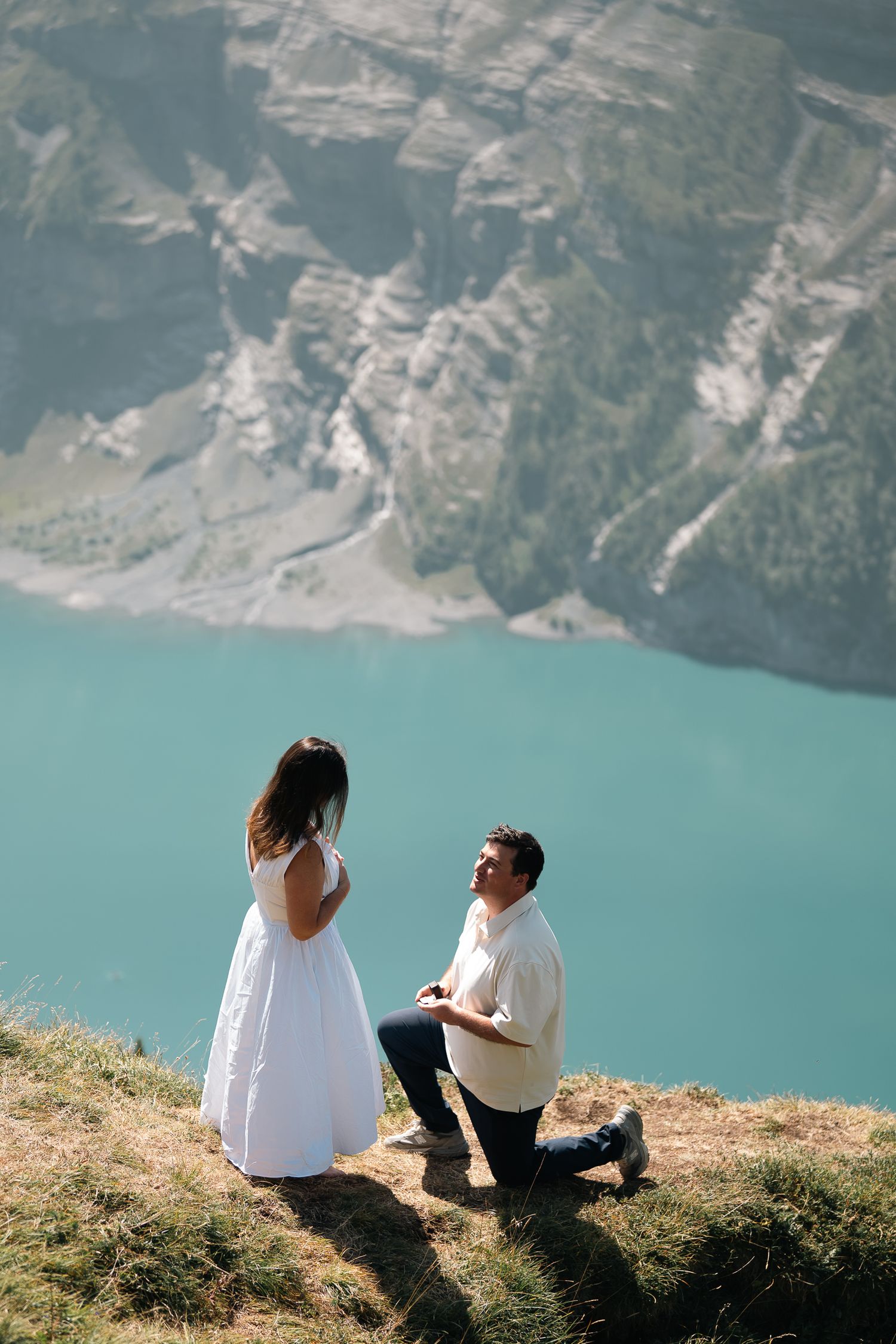Man proposes on one knee to woman in white dress on mountain cliff overlooking turquoise alpine lake.